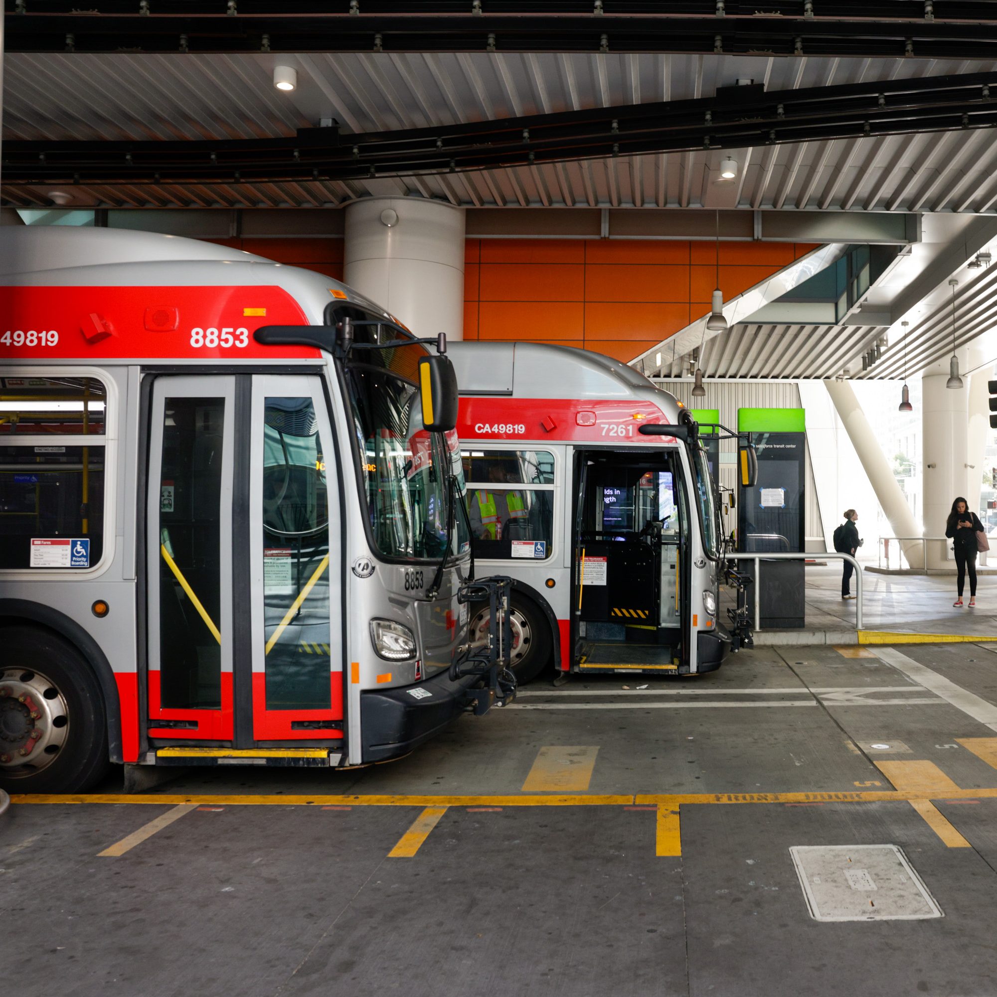 The image shows city buses parked in a well-lit, modern bus terminal with orange accents. Two people are seen walking nearby, and one bus has its doors open.