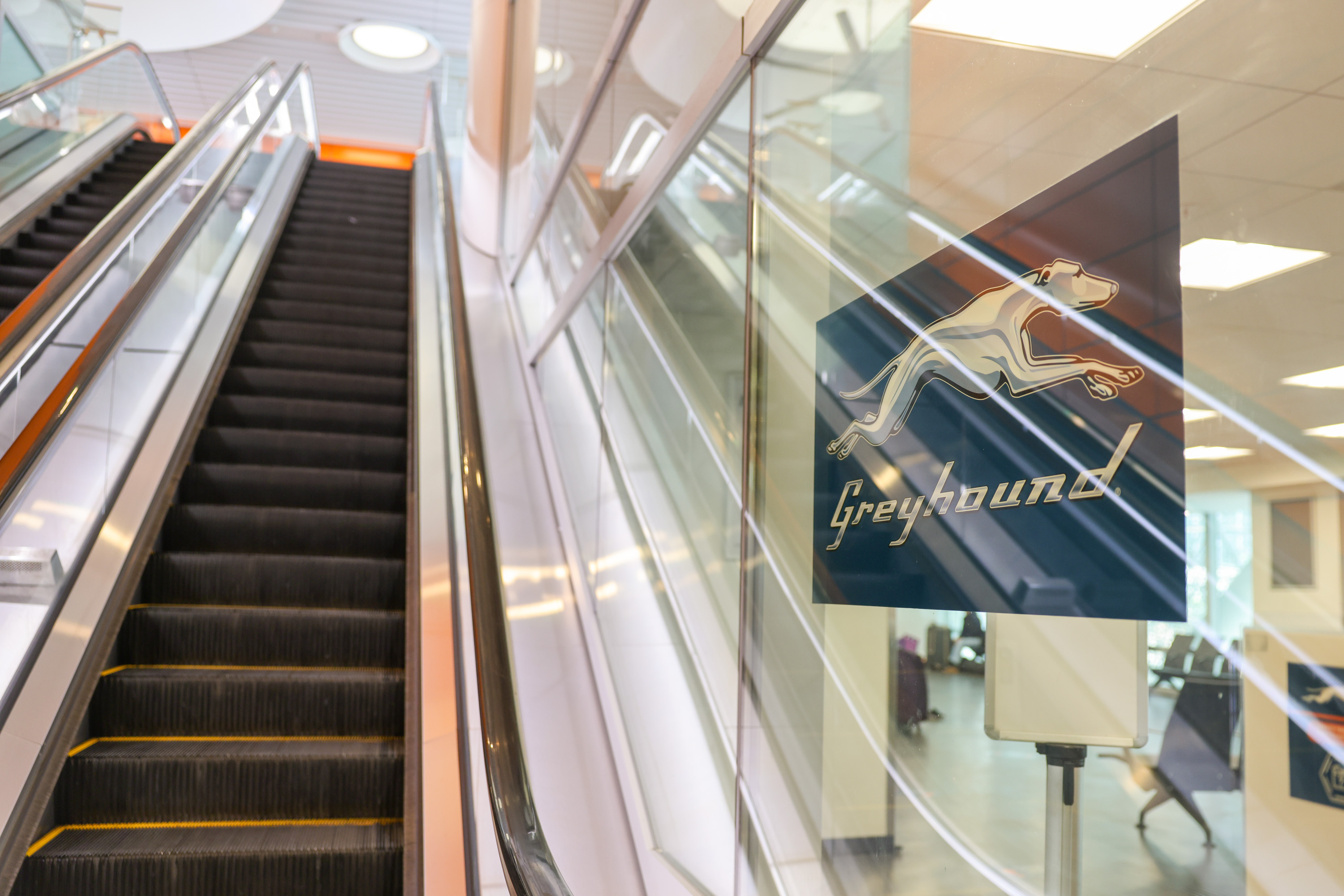 The image shows an escalator and a glass wall with a &quot;Greyhound&quot; logo, displaying a running greyhound. The background reveals an indoor area with seating.