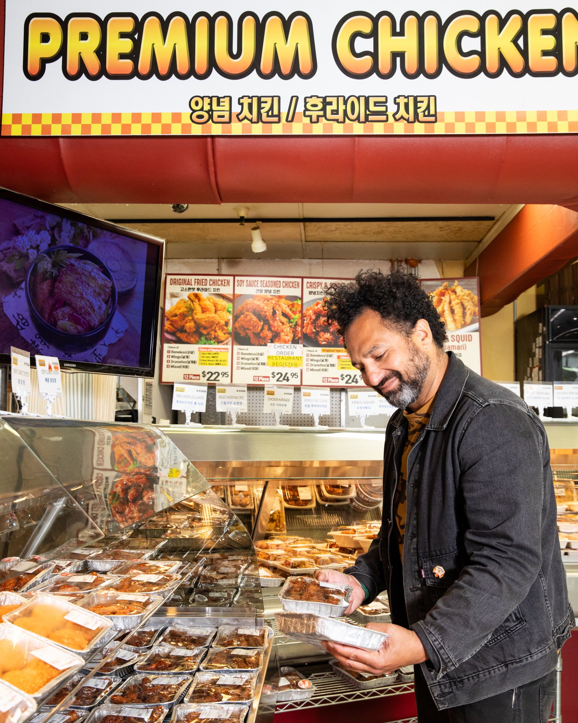 A man in a black jacket selects packaged food at a counter labeled “Premium Chicken,” with various fried chicken options displayed, and a screen shows a dish image.