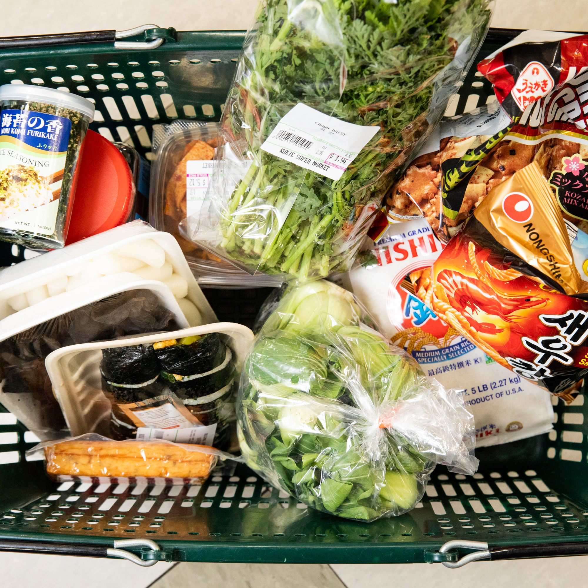 A grocery basket contains fresh greens, packaged sushi, Asian seasonings, noodles, and snacks, with a mix of fresh produce and ready-to-eat items.