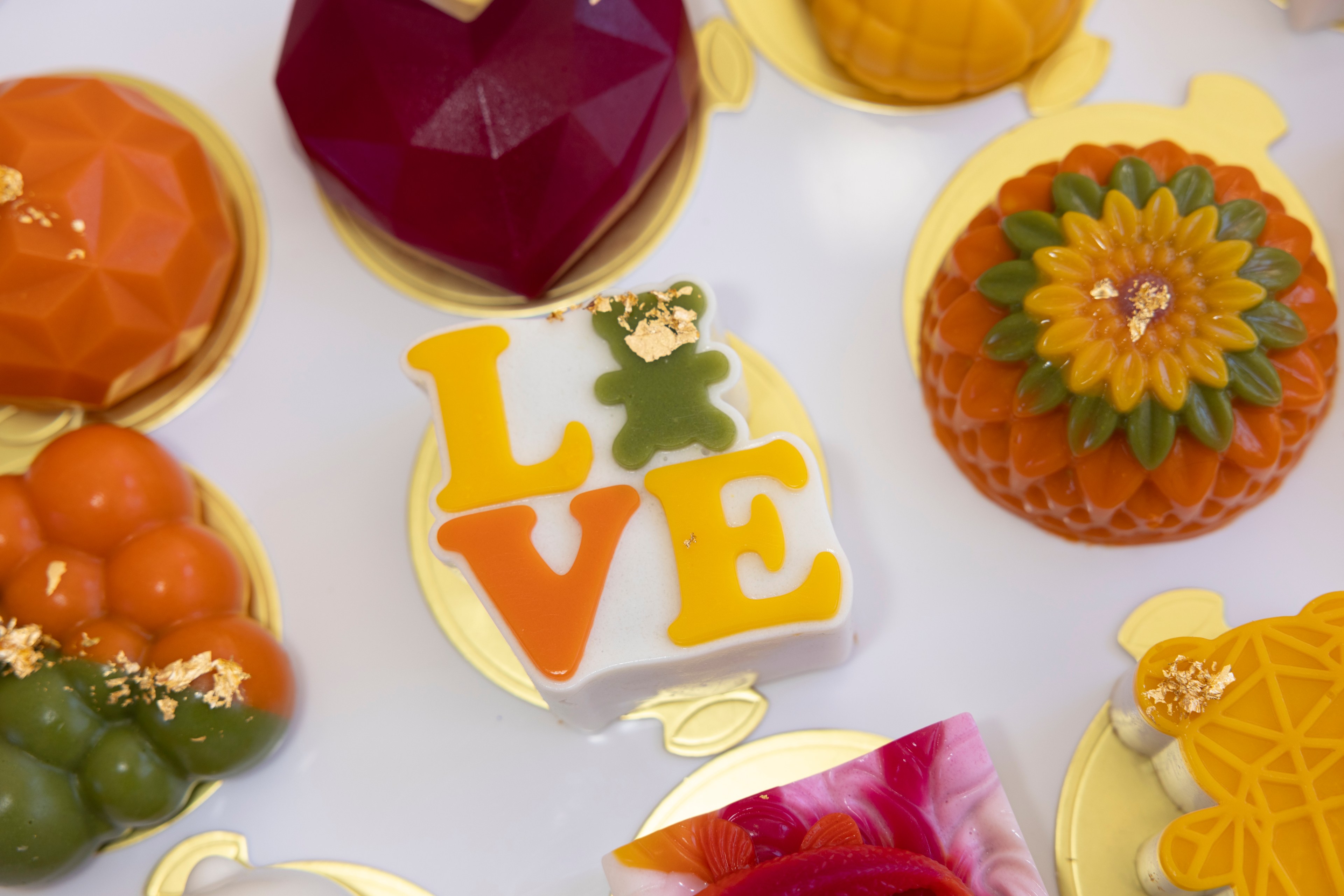 The image shows colorful, decorative pastries on gold trays, including a white square with &quot;LOVE&quot; in yellow and orange letters and a green teddy bear embellishment.