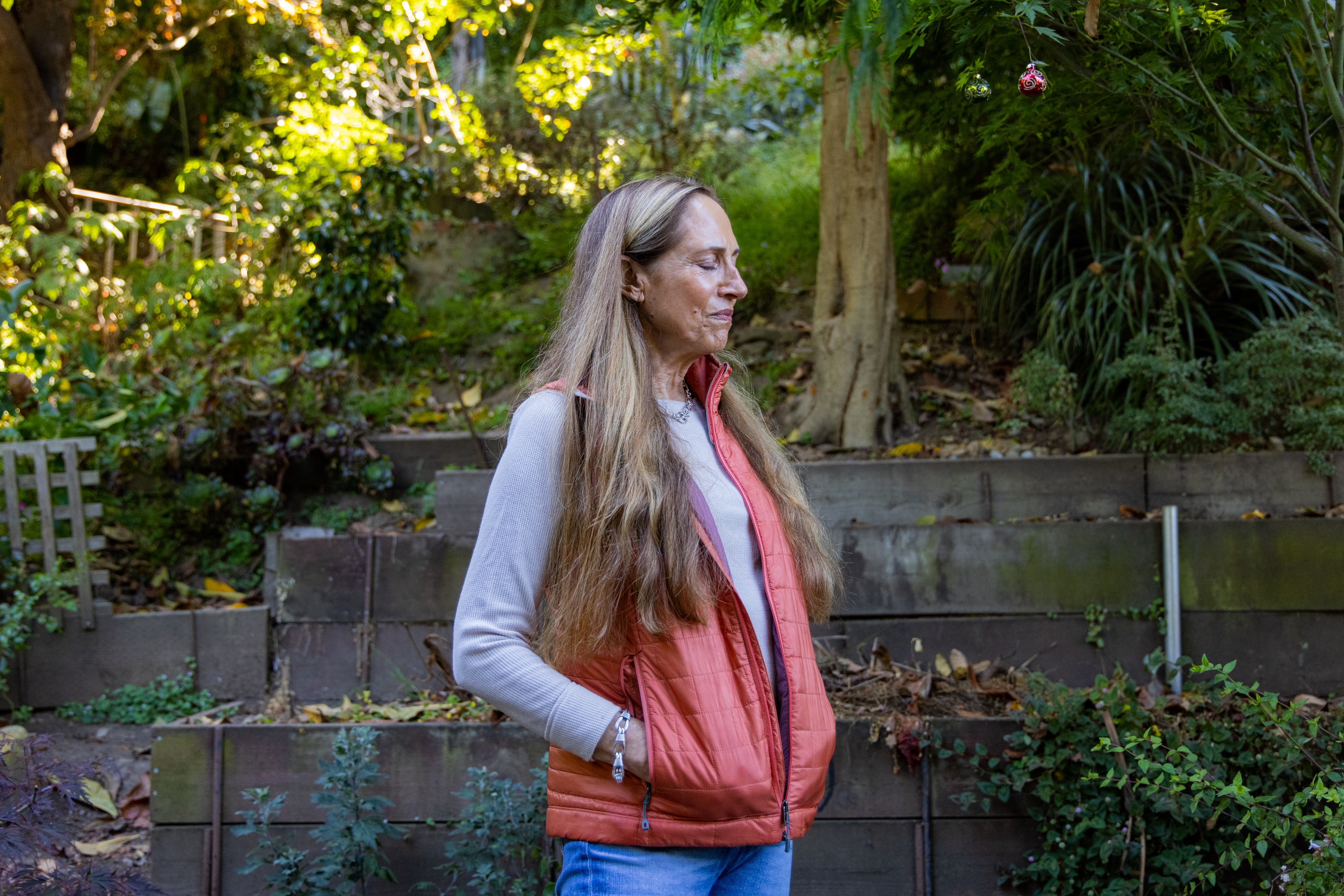 A person with long hair in a red vest stands in a garden with lush greenery, tiered stone walls, and a large tree in the background.