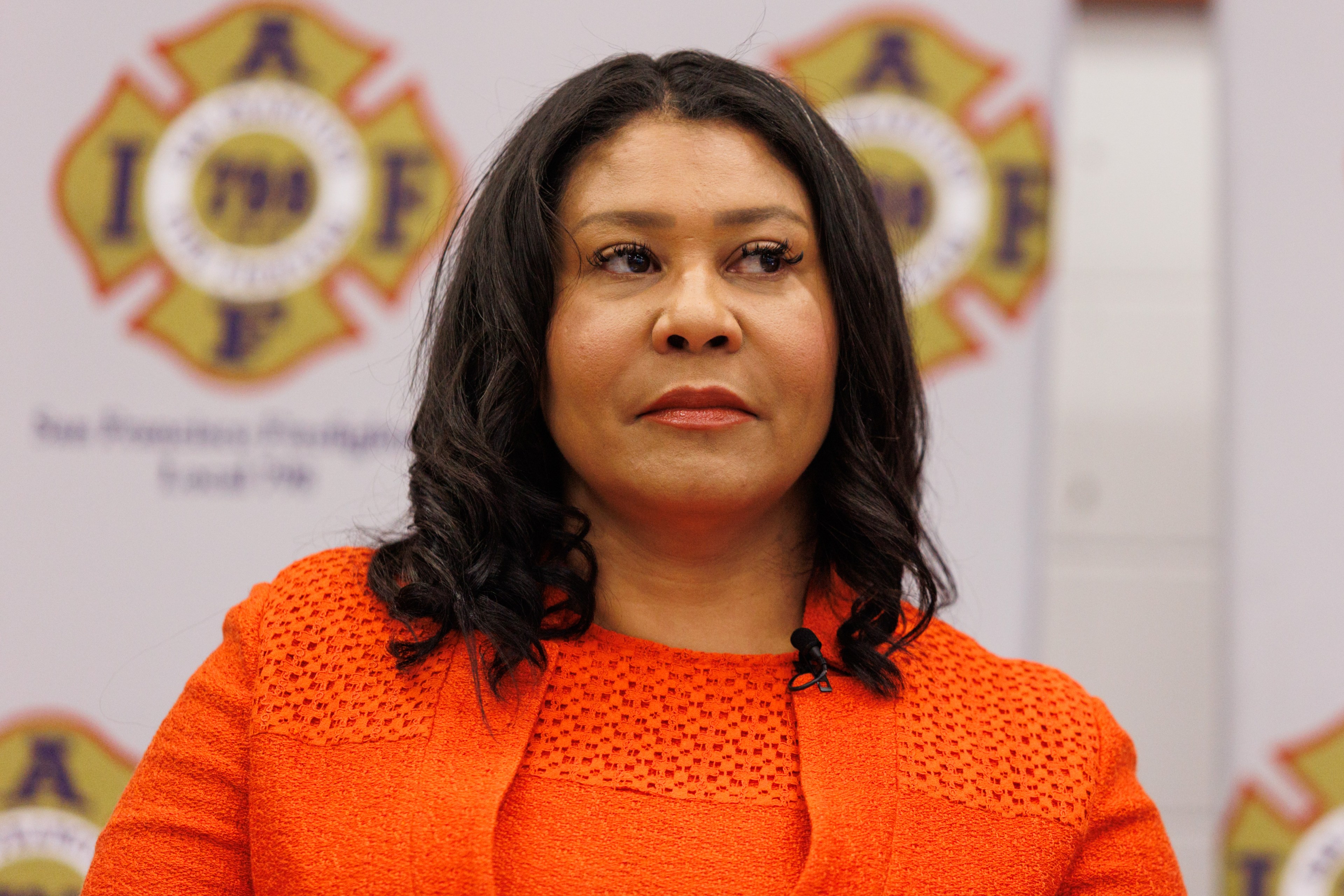 A woman with dark, wavy hair wears a bright orange top and stands against a backdrop featuring fire department emblems. Her expression is serious.