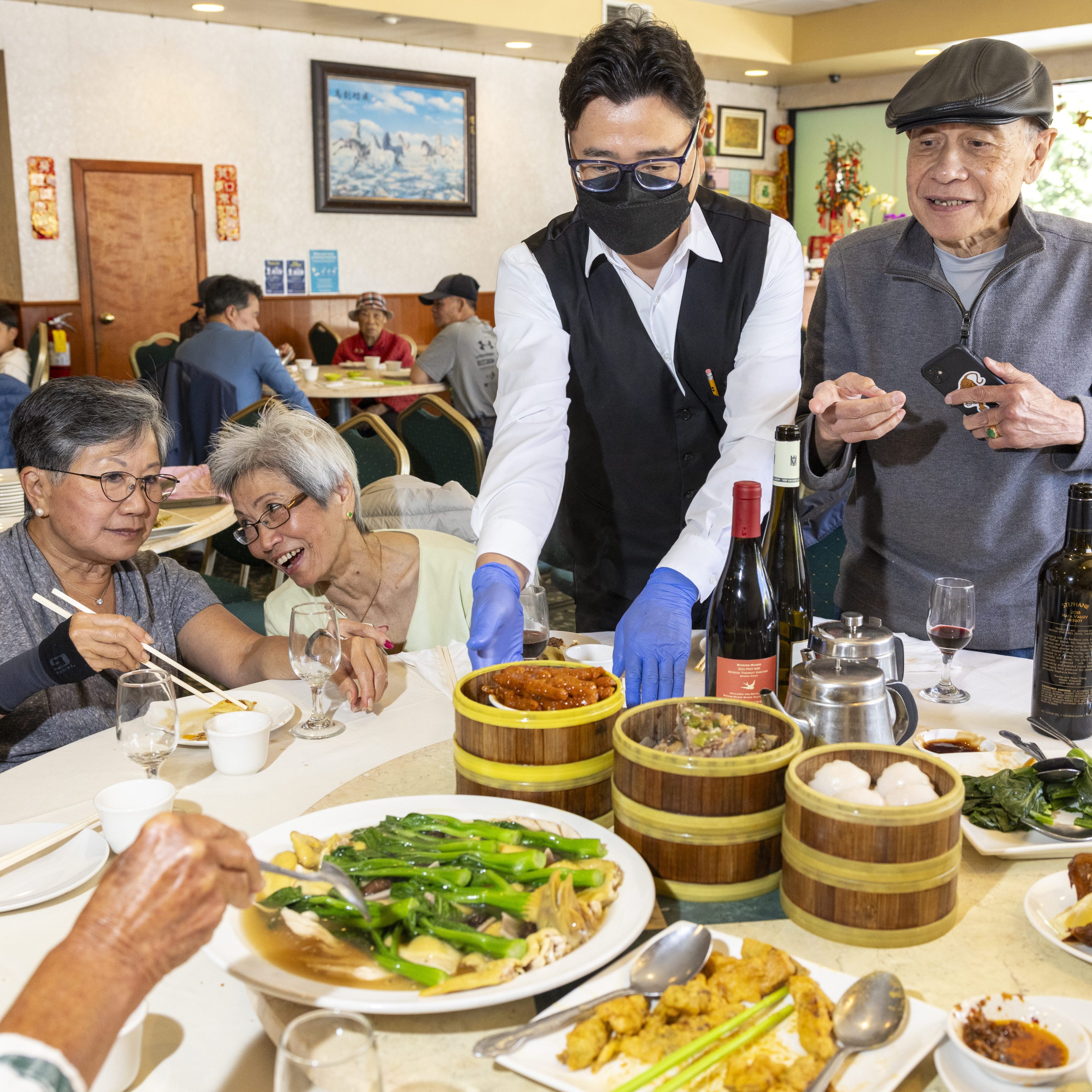 An attentive waiter in a black vest and gloves serves food to a cheerful group of diners in a well-decorated restaurant, with dim sum and wine on the table.