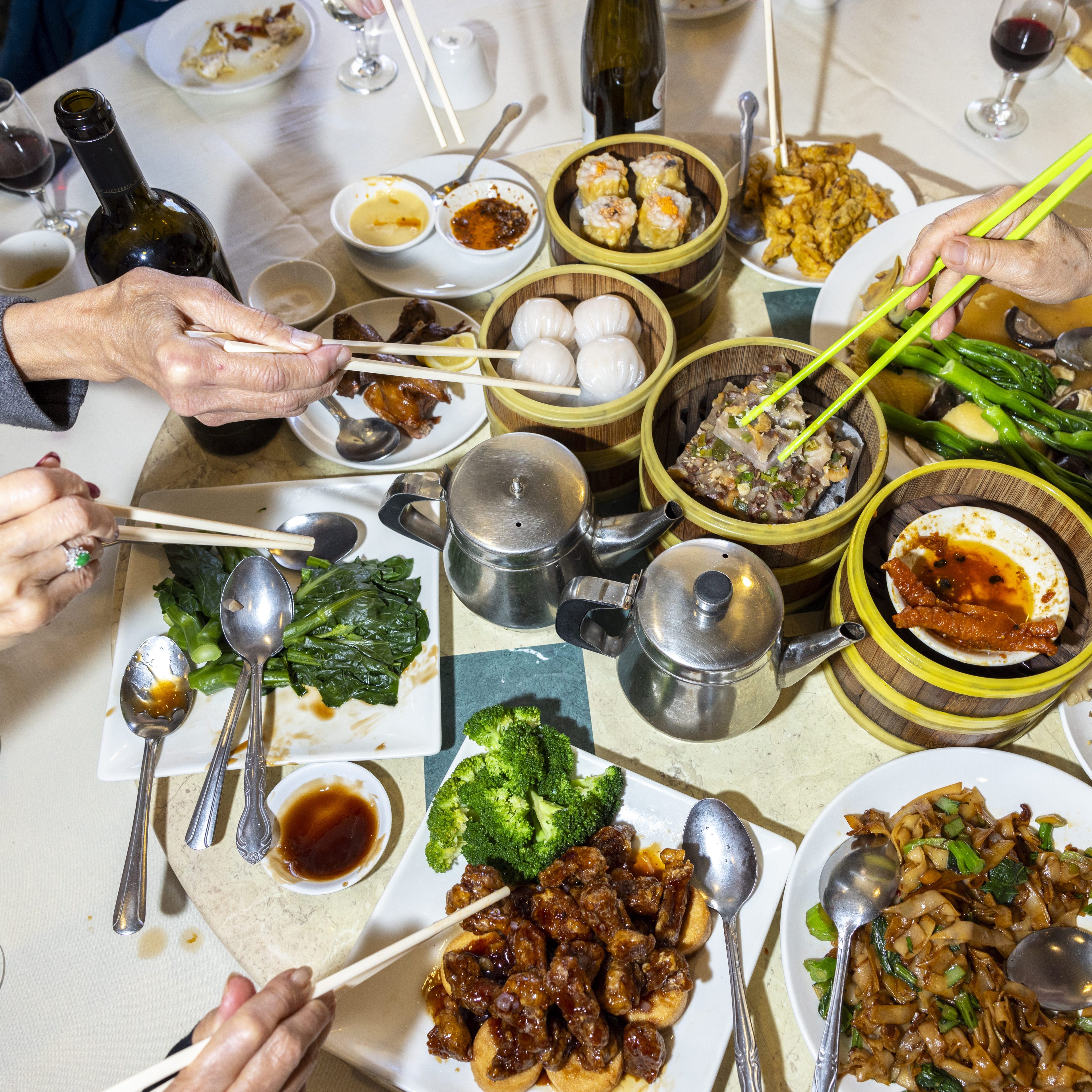 A table is filled with various Chinese dishes, including dim sum, noodles, broccoli, and greens, with several hands using chopsticks to serve themselves.