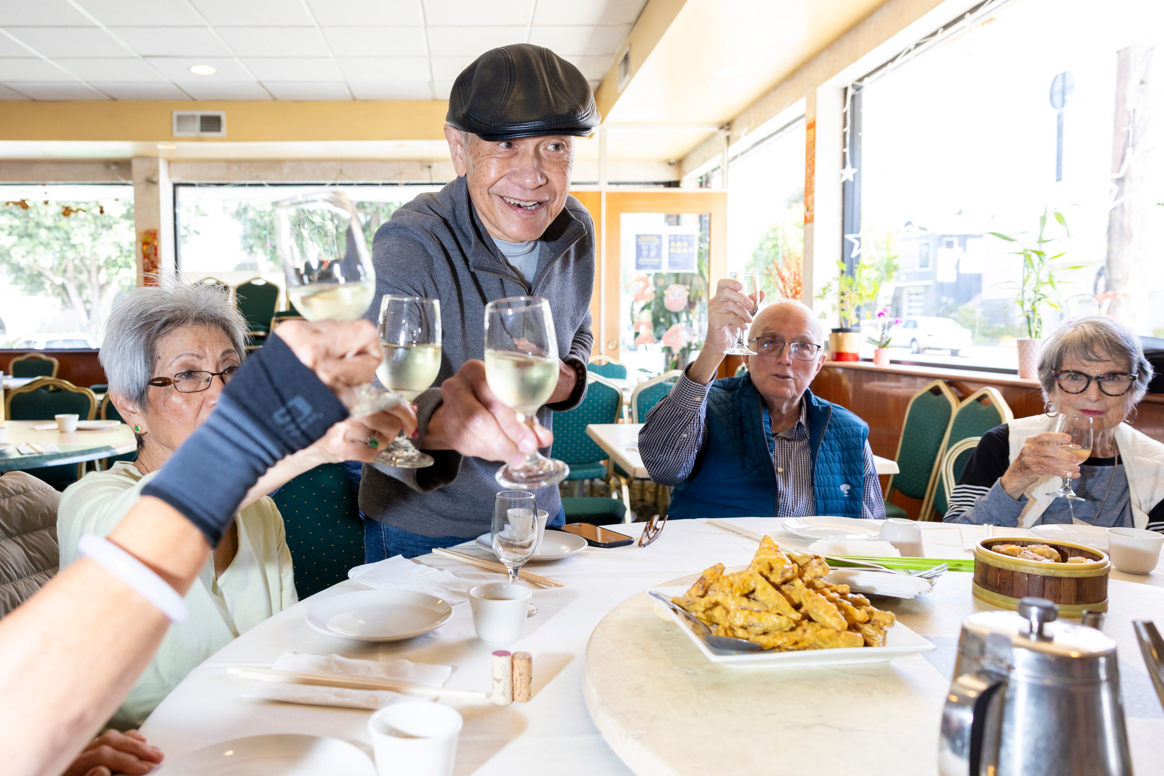 Five elderly friends are seated around a restaurant table, toasting with glasses of white wine. The table is set with food and dishes, and the mood is jubilant.