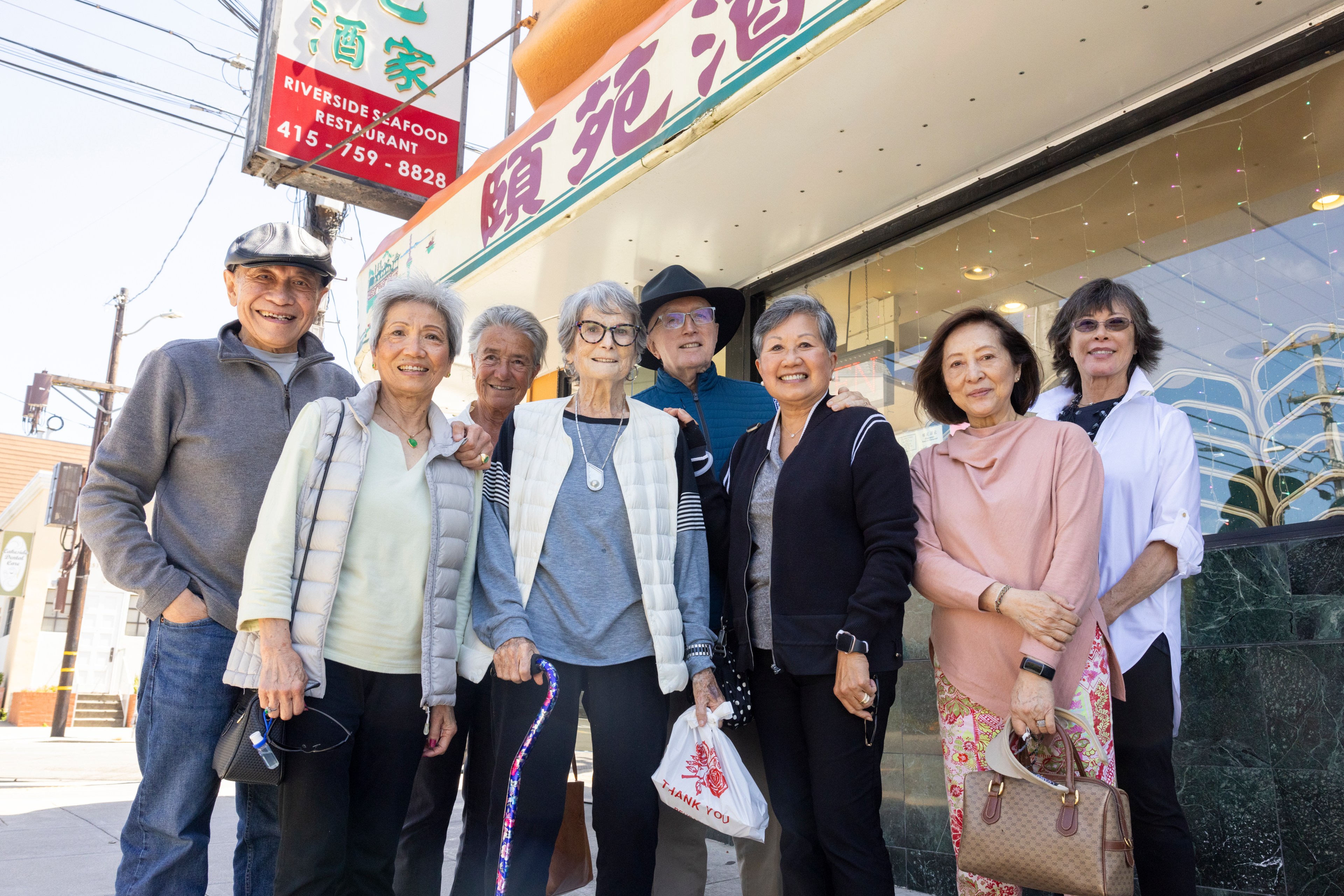 A group of eight smiling elderly individuals stands outside a Riverside Seafood Restaurant, posing for a photo. They seem happy and sociable, dressed casually.