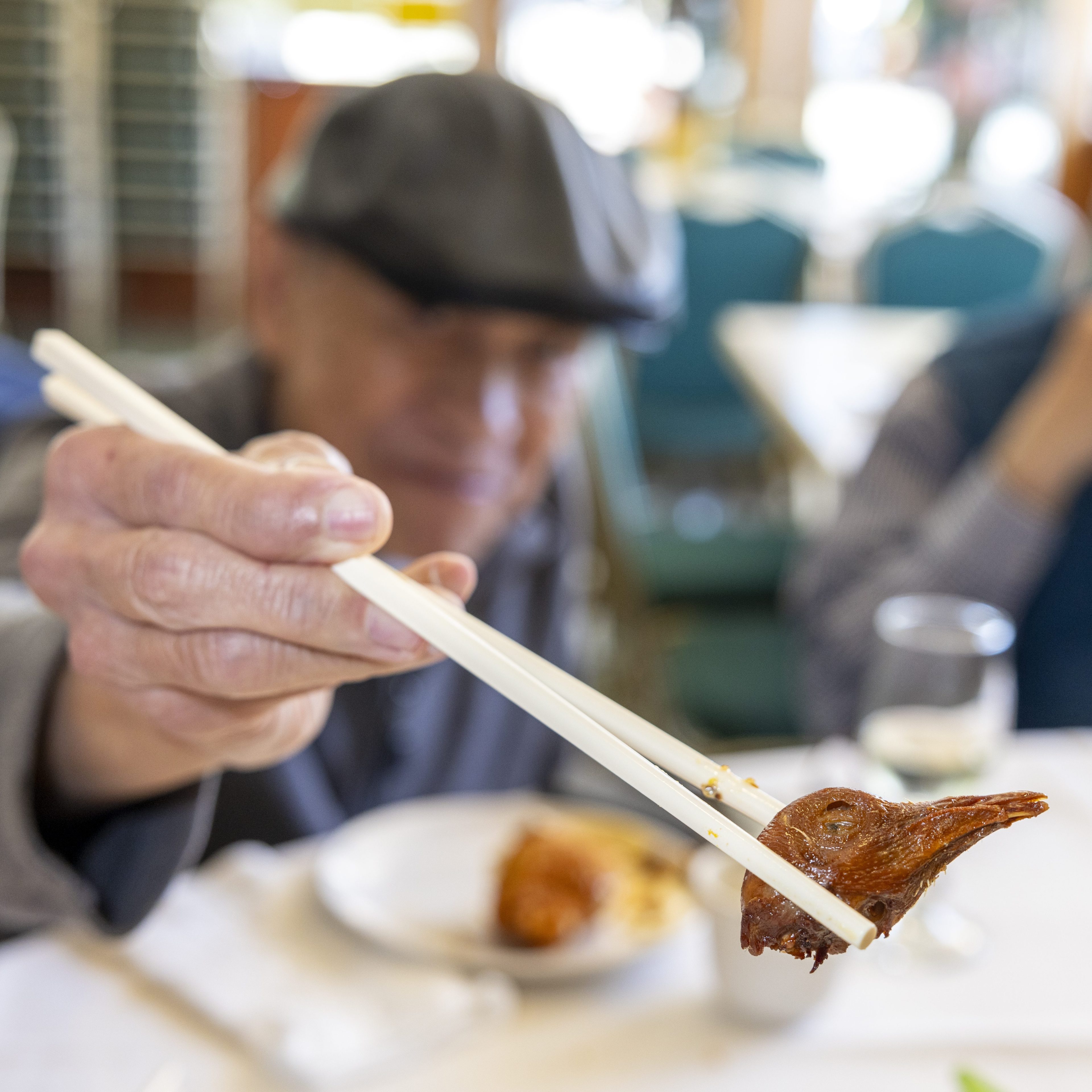 An elderly person in a hat is holding a cooked pigeon head with chopsticks, sitting at a table in a restaurant with blurred background.