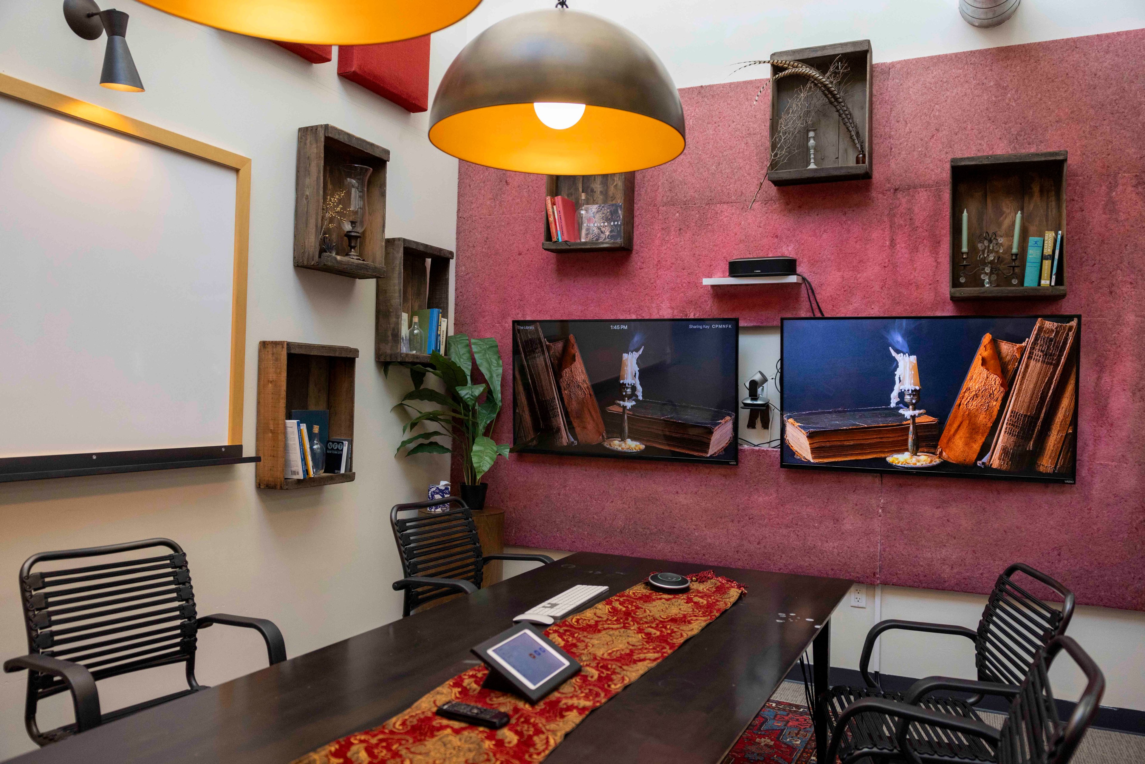 The image shows a cozy conference room with a wooden table, black chairs, wall-mounted shelves, two TVs, a plant, and large ceiling lamps.