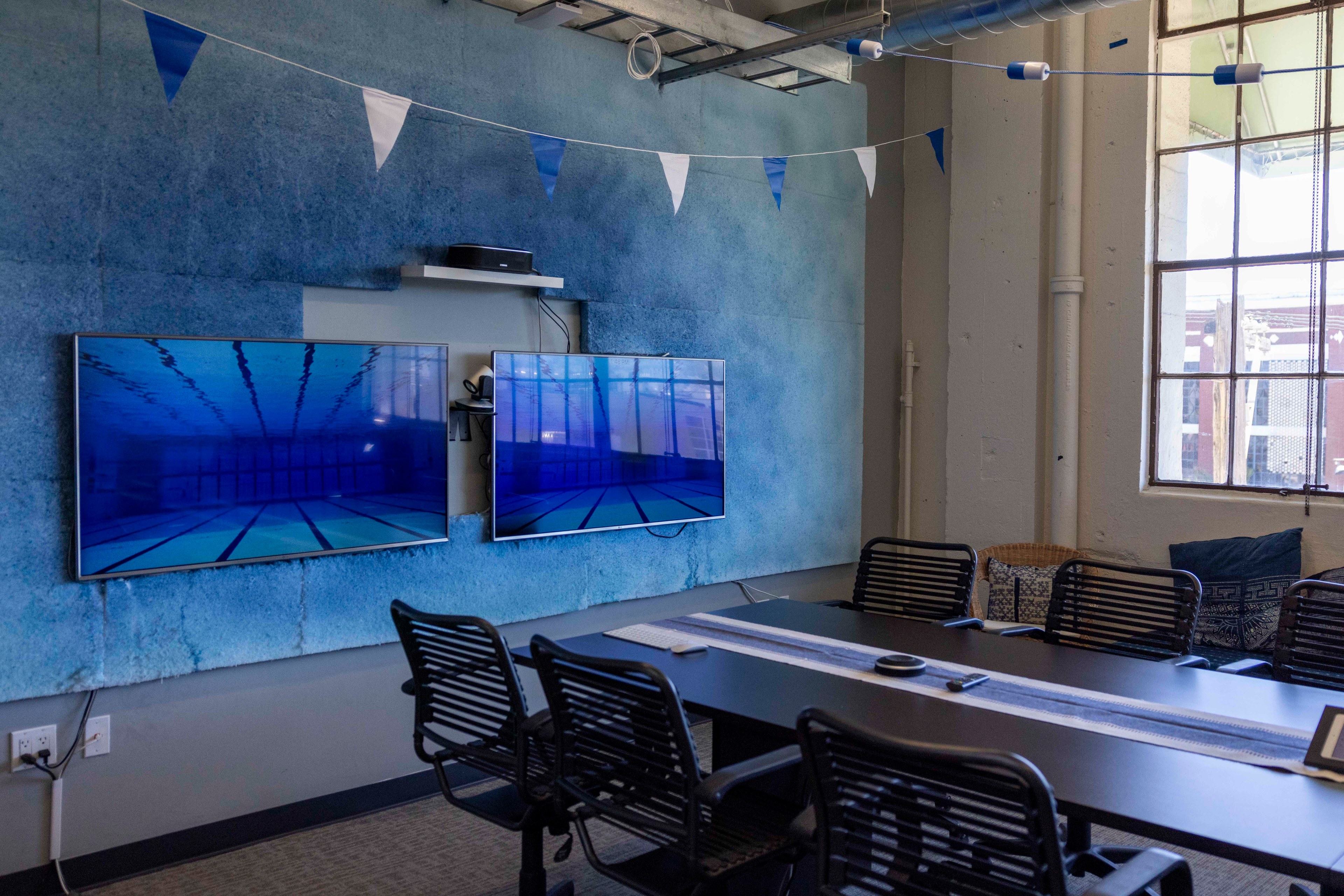 A conference room with six black chairs around a rectangular table, two wall-mounted screens displaying underwater pool scenes, and blue-white bunting overhead.