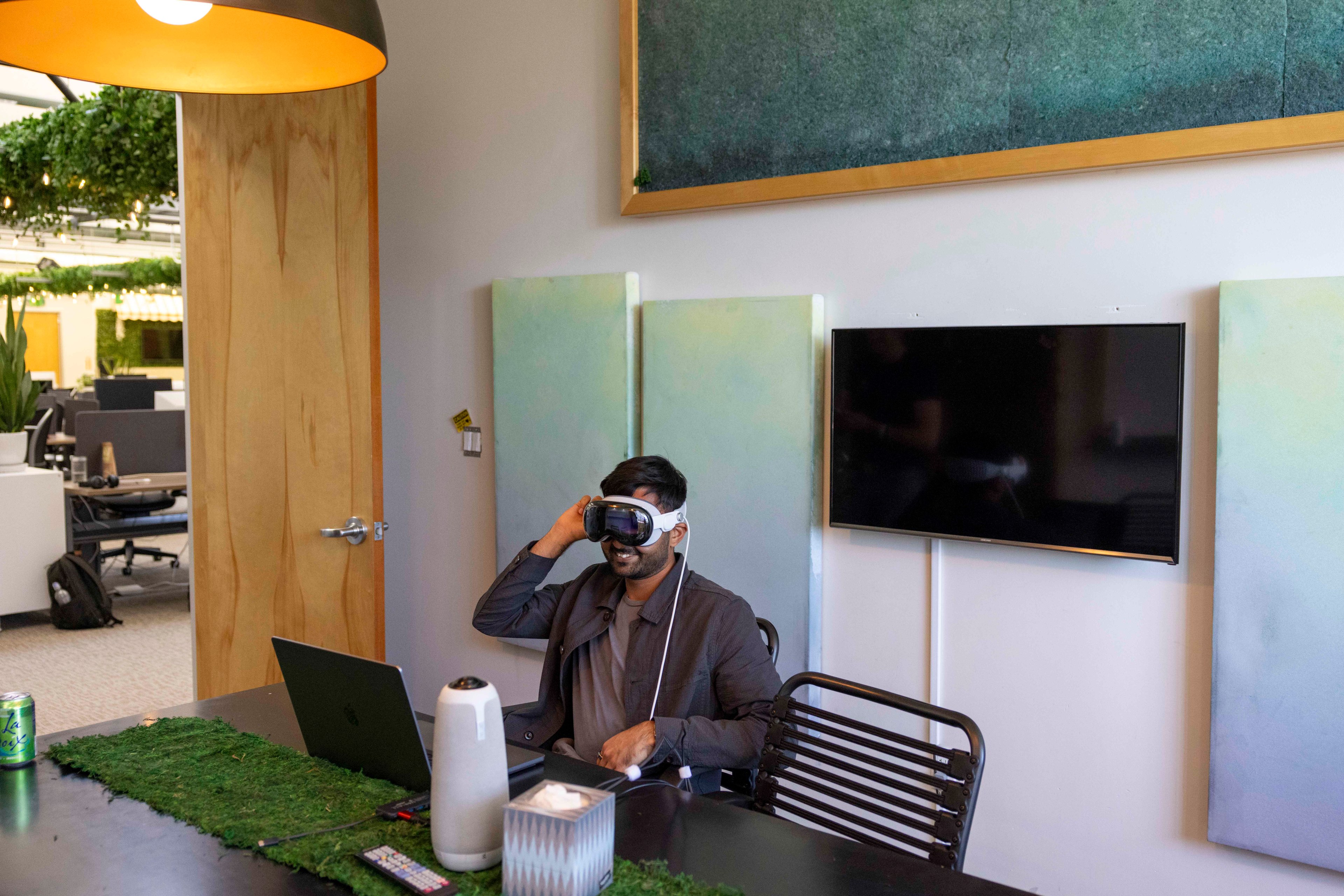 A person wearing VR goggles sits at a desk with a laptop in a modern, plant-decorated office, featuring a large TV and soft green color panels on the wall.