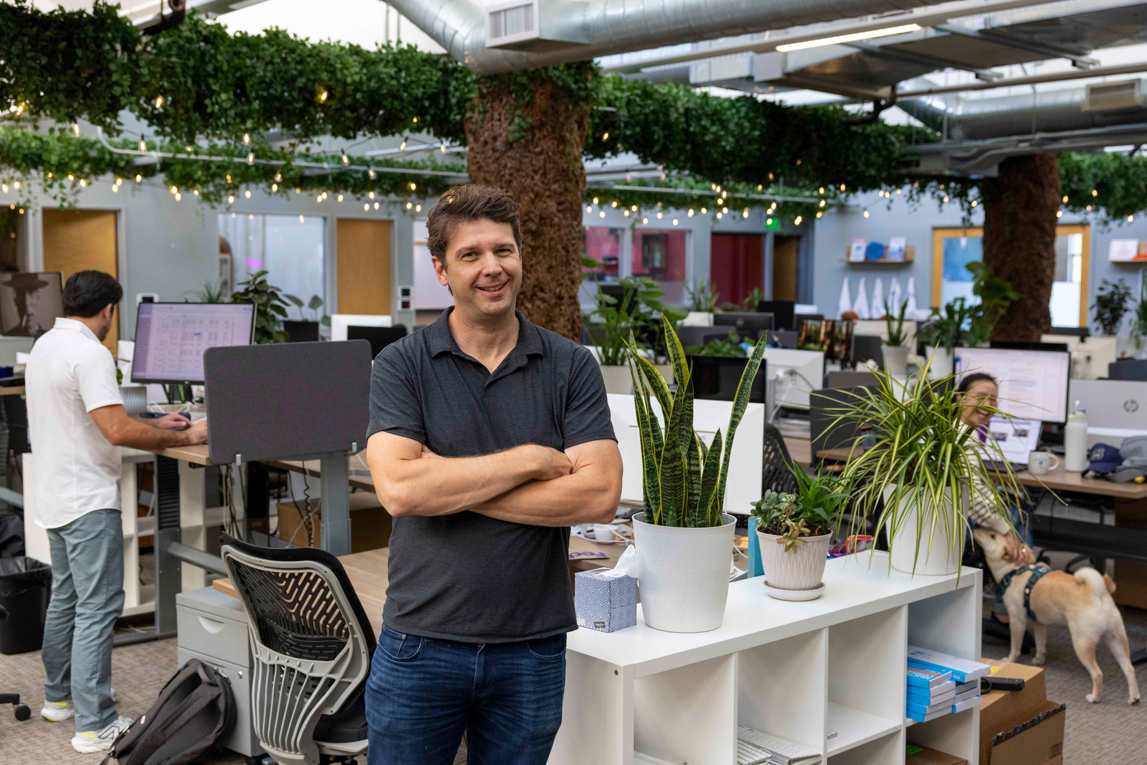 A man stands smiling in an office with arms crossed, surrounded by plants and colleagues working at desks. Greenery decorates the area; a dog is present.