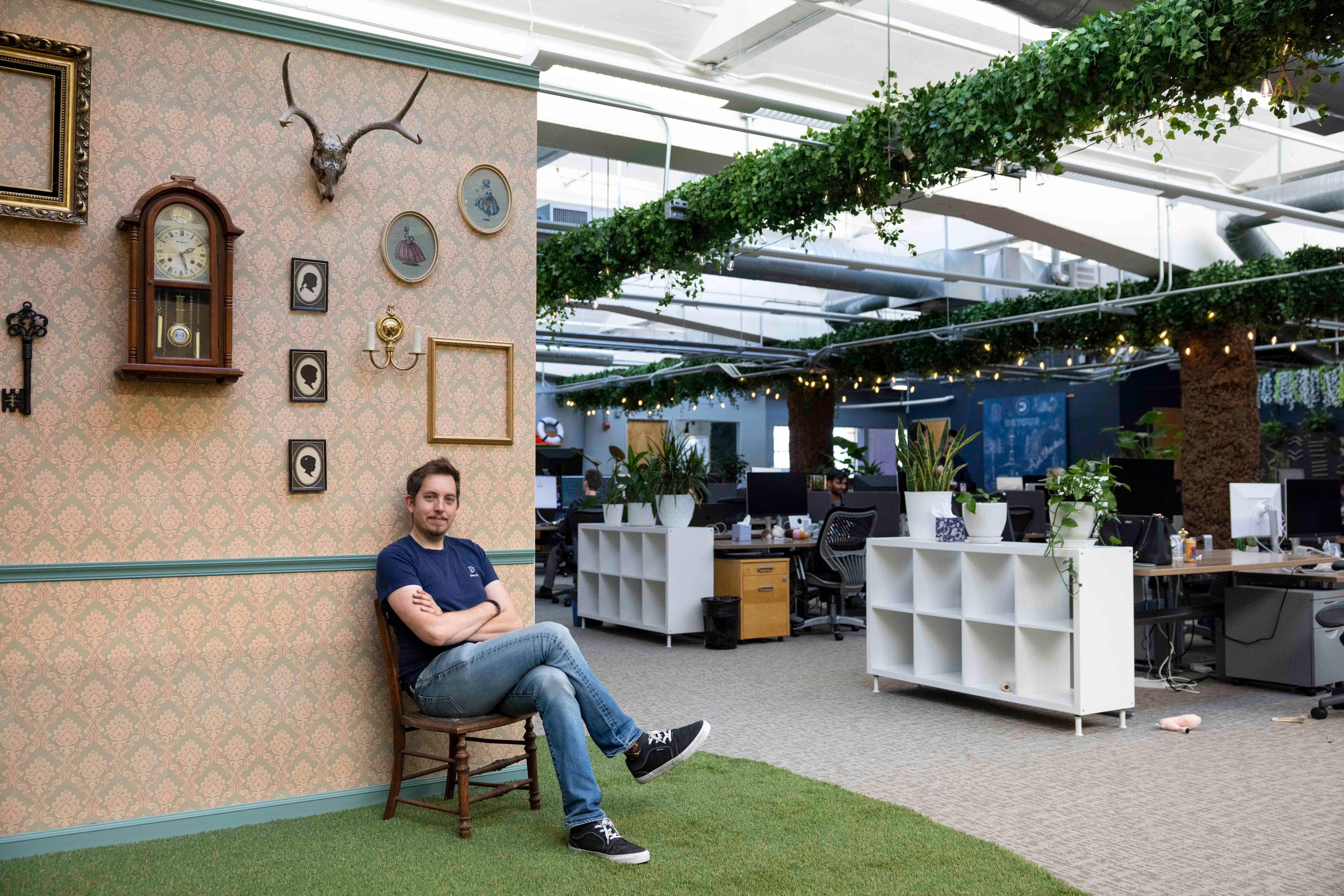 A man in a casual outfit sits on a chair against a decorated wall in a spacious, modern office with plants and fairy lights hanging from the ceiling.