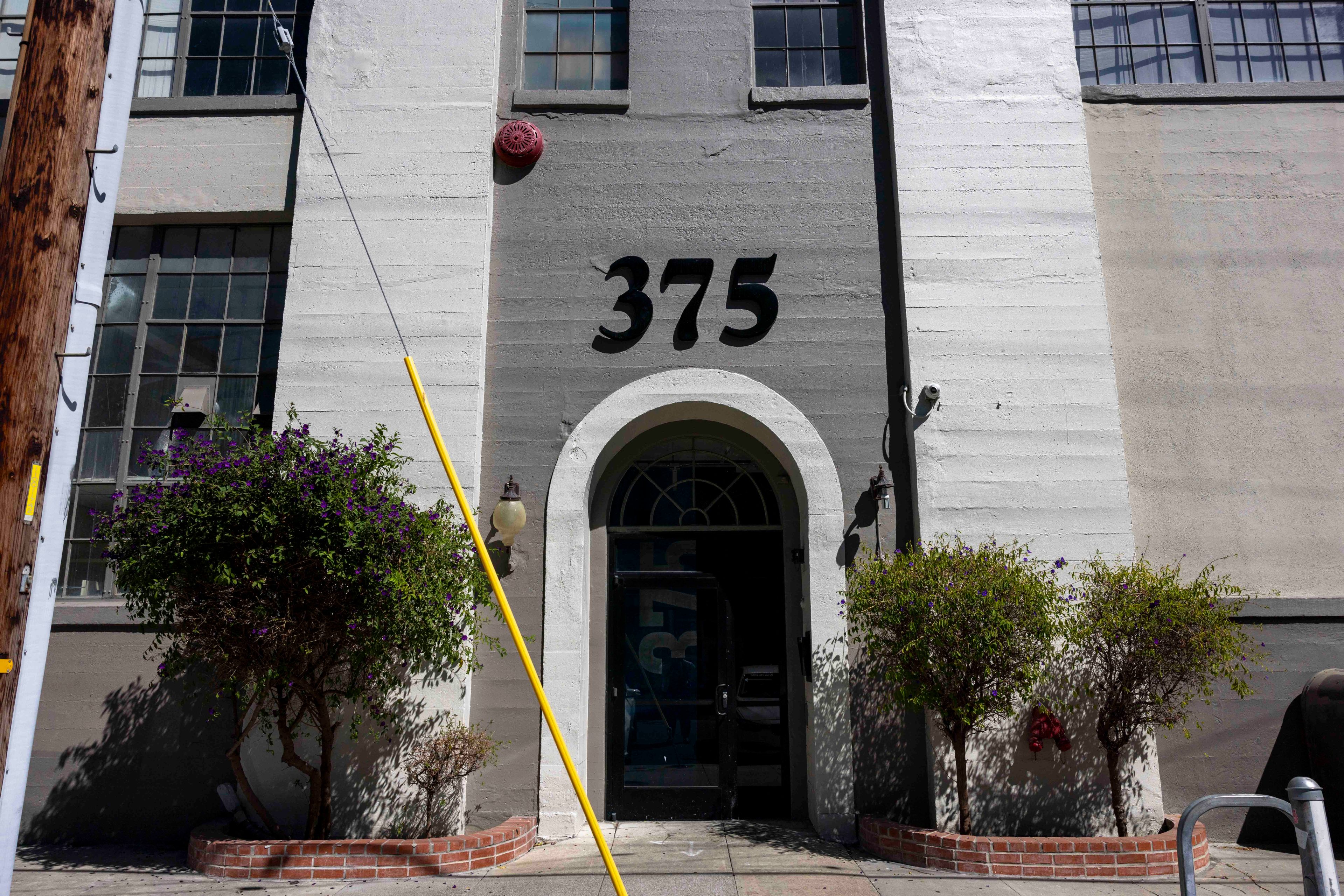 The image shows a building with the number 375 above an arched entrance. Two small trees stand on either side of the doorway, which is flanked by two large windows.