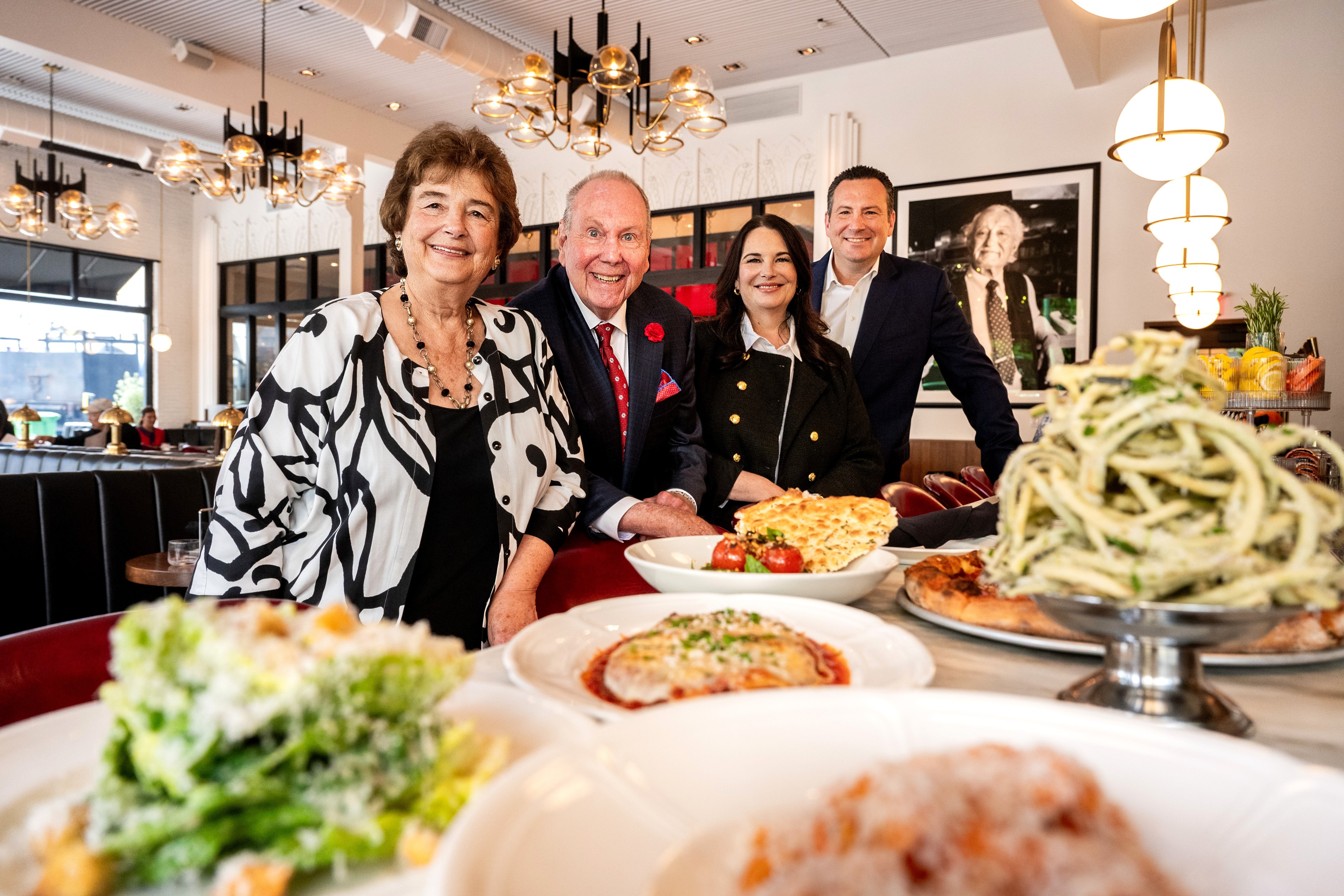 Four people in a well-lit restaurant smile at the camera, with plates of various dishes in the foreground, including pasta, pizza, and salad.