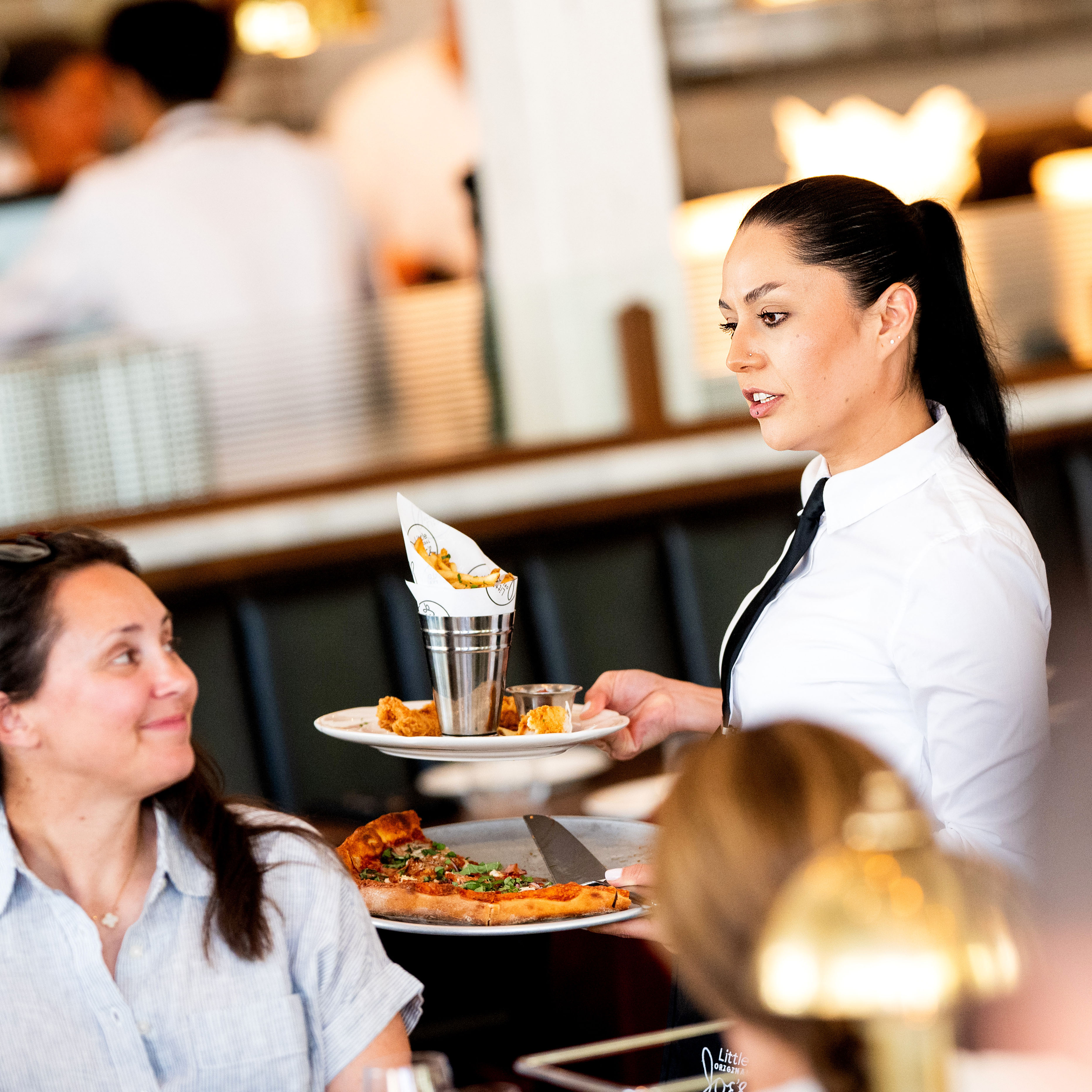 A waitress in a white shirt and black tie serves a smiling woman a pizza and another dish in a busy, well-lit restaurant.