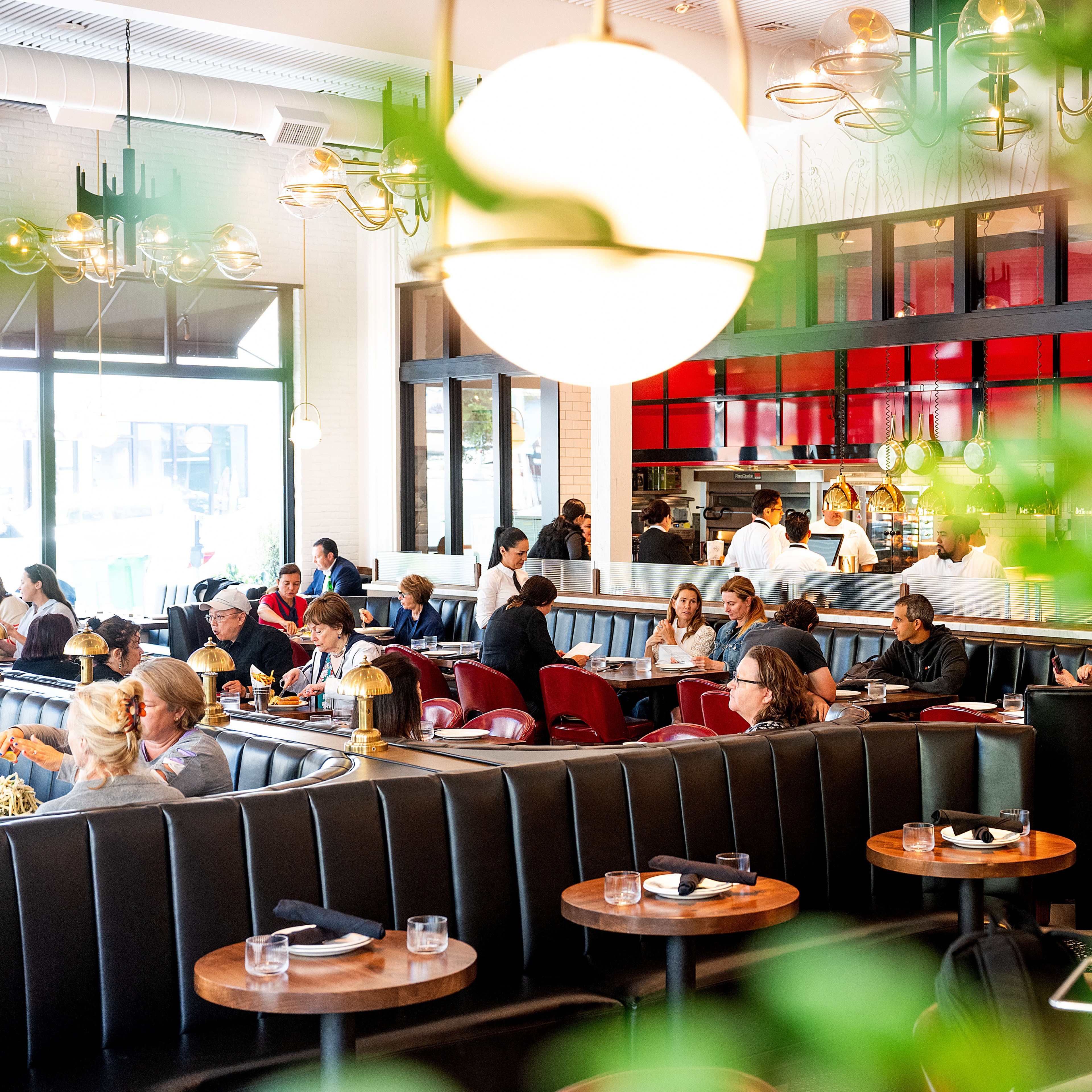 A busy, modern restaurant with red and black decor, people dining at tables and booths, bright lighting, and an open kitchen in the background.