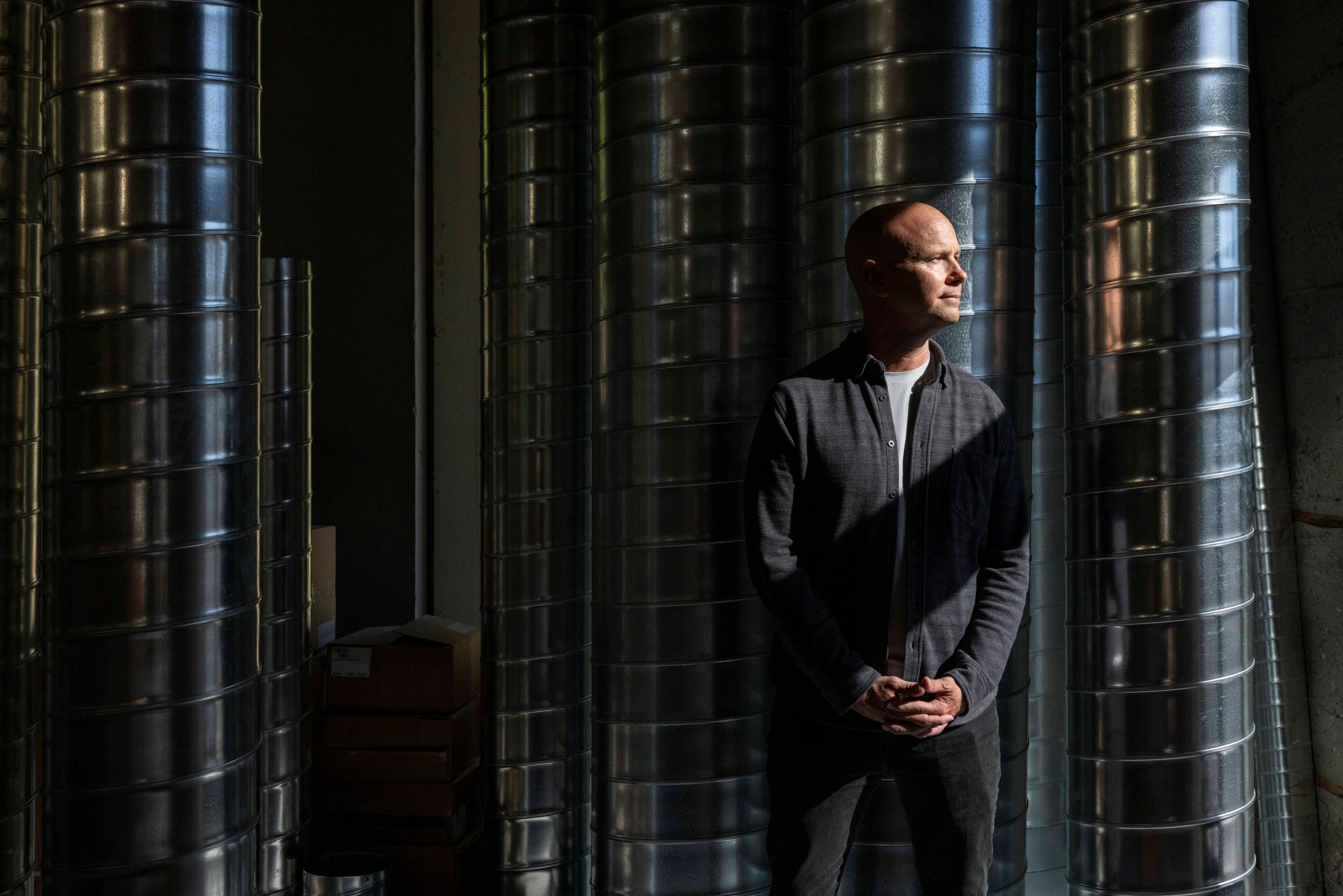 A person stands in a dimly lit room surrounded by large metal ducts. They are dressed in a dark shirt and pants, with light casting shadows across their face.