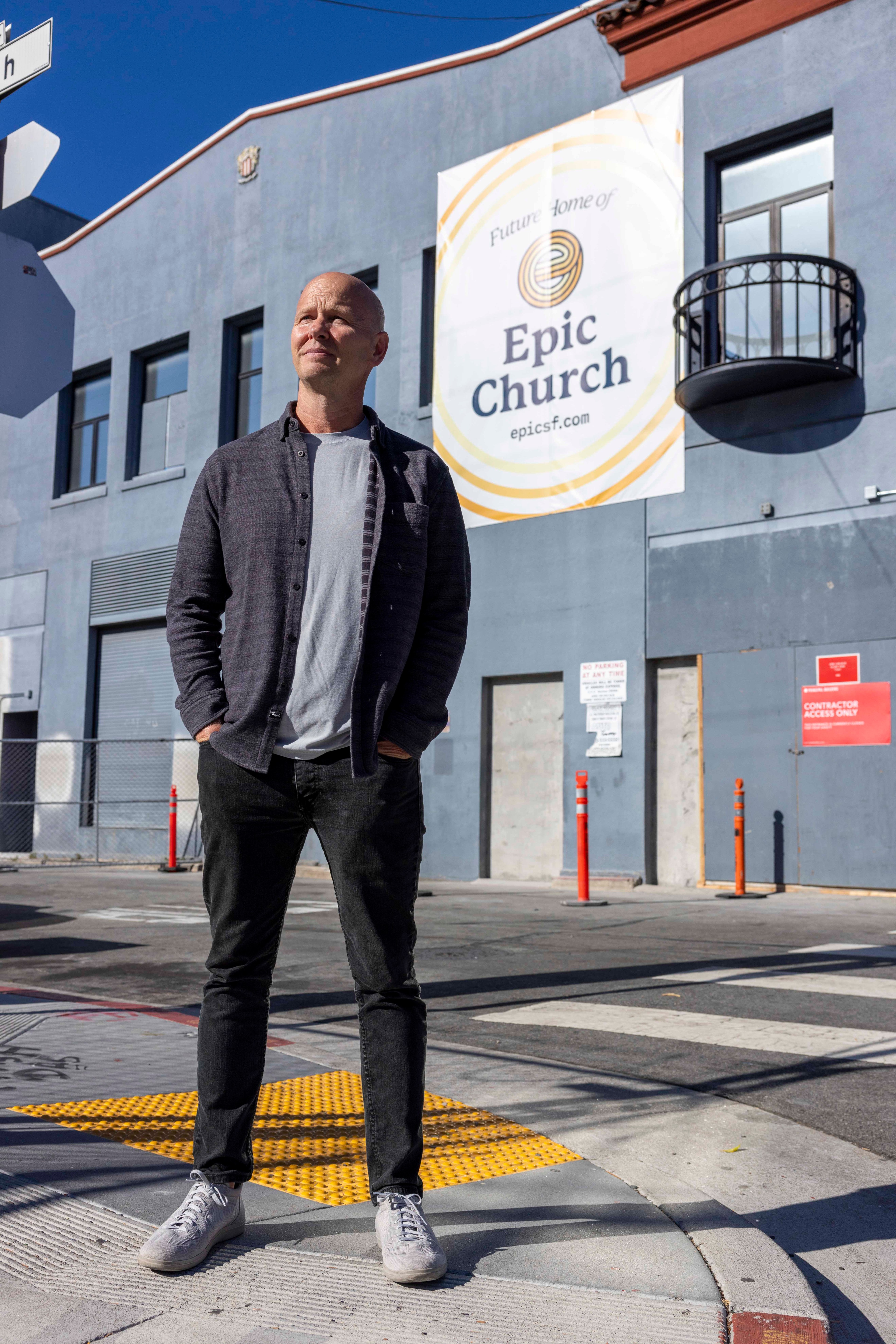 A person stands on a street corner in front of a blue building with a sign reading "Future Home of Epic Church" and looks thoughtfully into the distance.