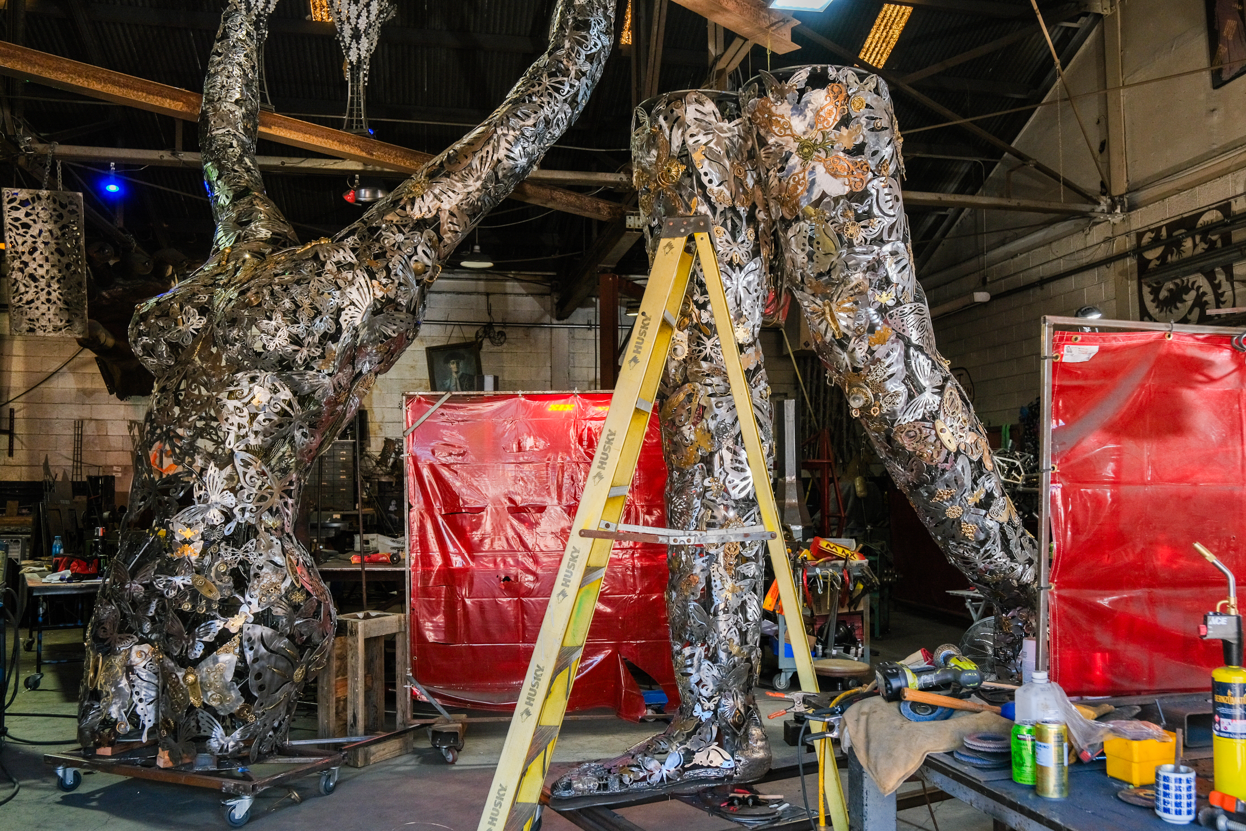 The image depicts a workspace with large, intricate metal sculptures in progress, featuring a human form. A yellow ladder and various tools are also visible in the busy workshop.