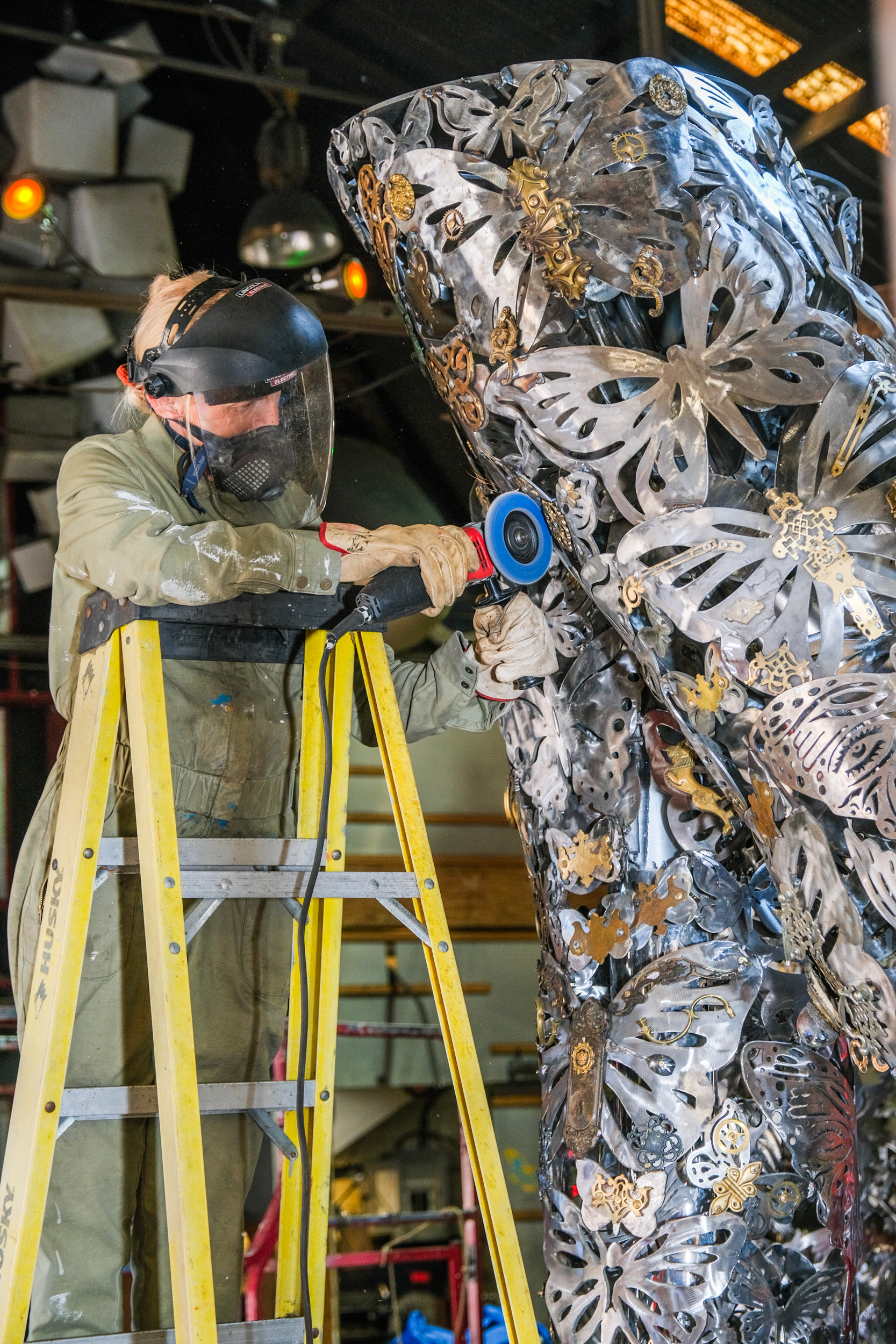 A person wearing safety gear stands on a yellow ladder, using a power tool to work on a large metal sculpture adorned with intricate butterfly designs.