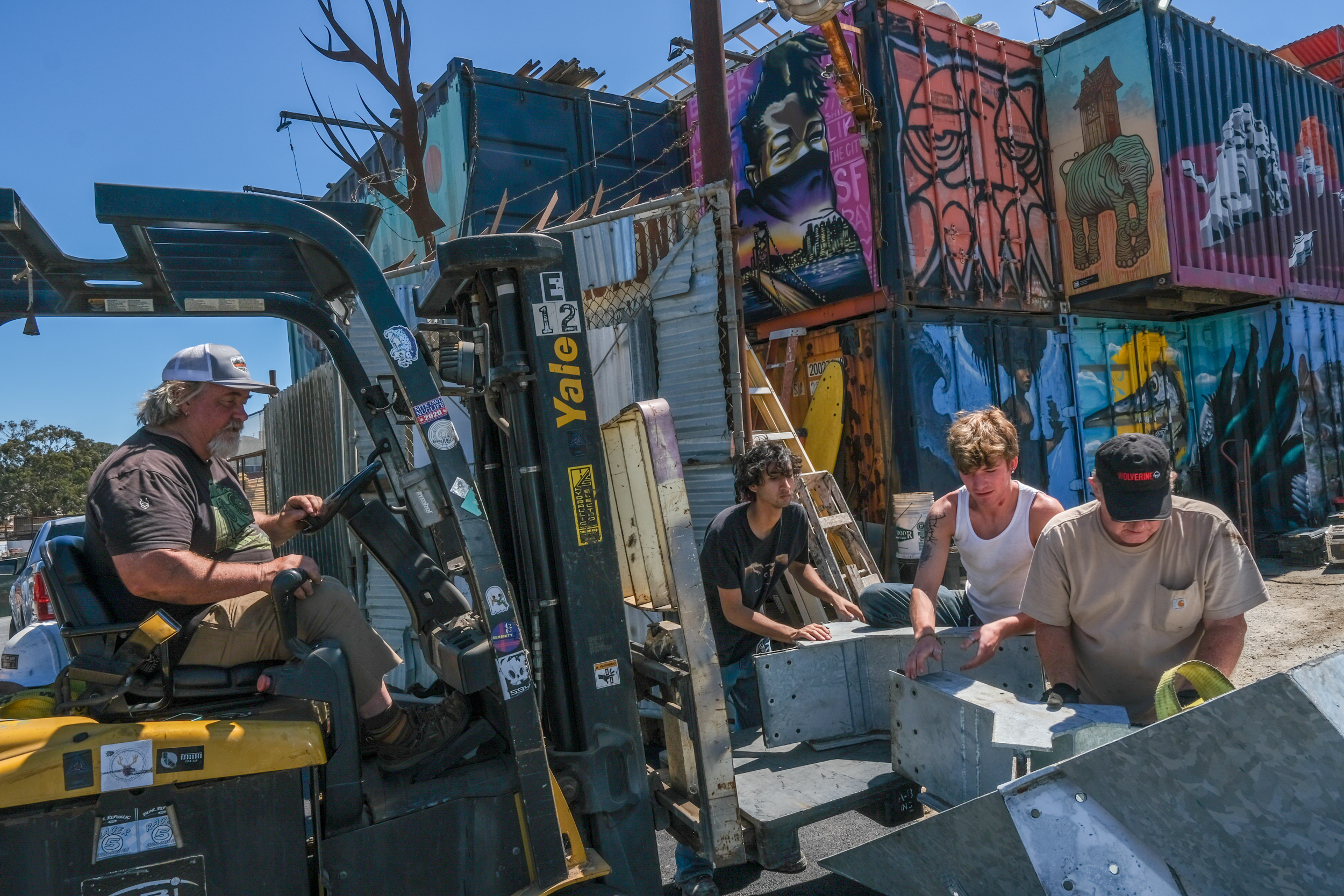 A man operates a forklift in front of several individuals handling metal parts. The background features vividly painted shipping containers.