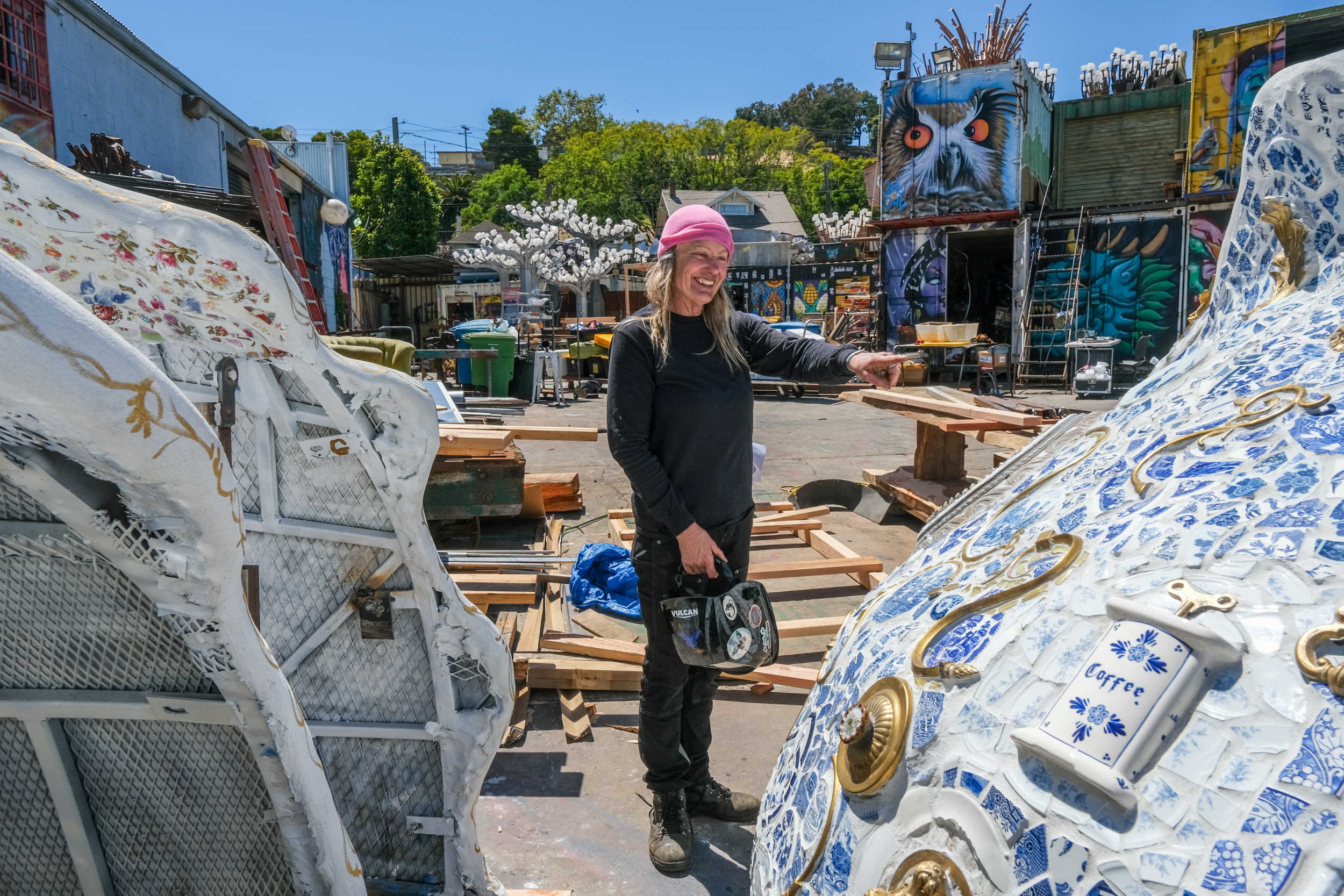 A person with long hair and a pink hat points while standing in an art-filled yard, surrounded by large, colorful sculptures, including a blue and white mosaic piece.