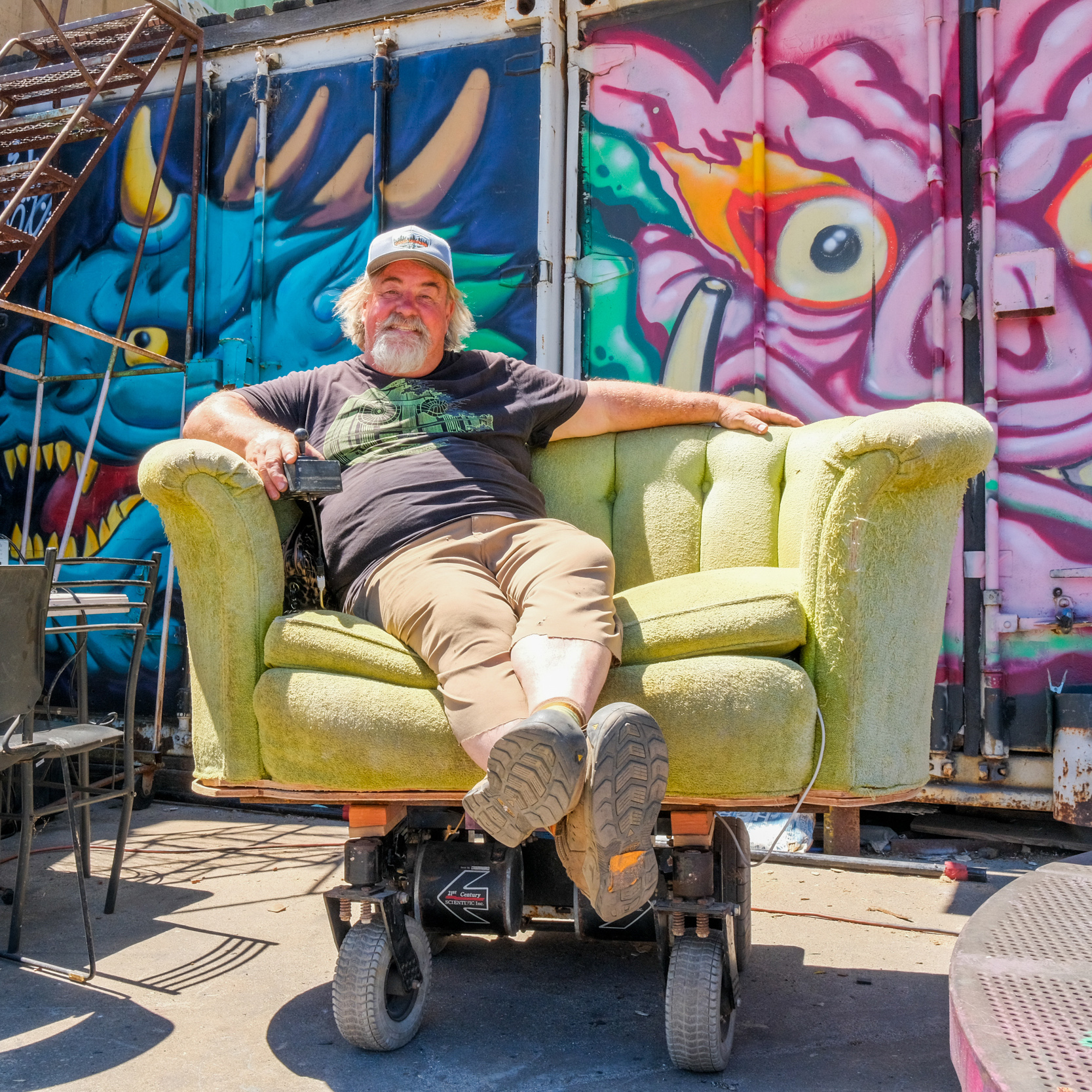 A man with a beard and cap sits on a light green chair with wheels, in front of a vibrant mural featuring colorful dragon and monster faces.