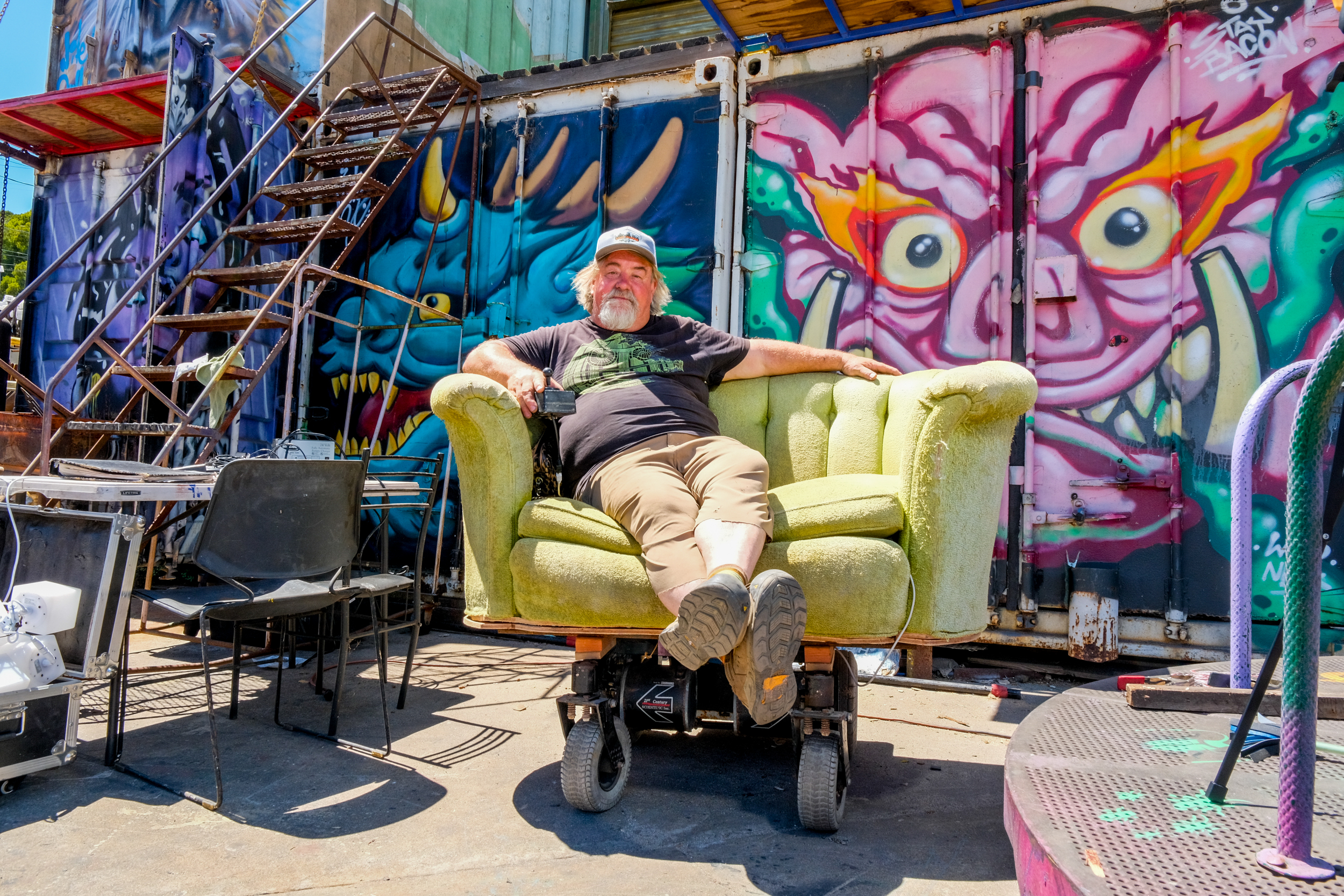 A man sits on a green upholstered chair outside, surrounded by colorful graffiti art featuring whimsical creatures, with metal stairs and scattered chairs nearby.