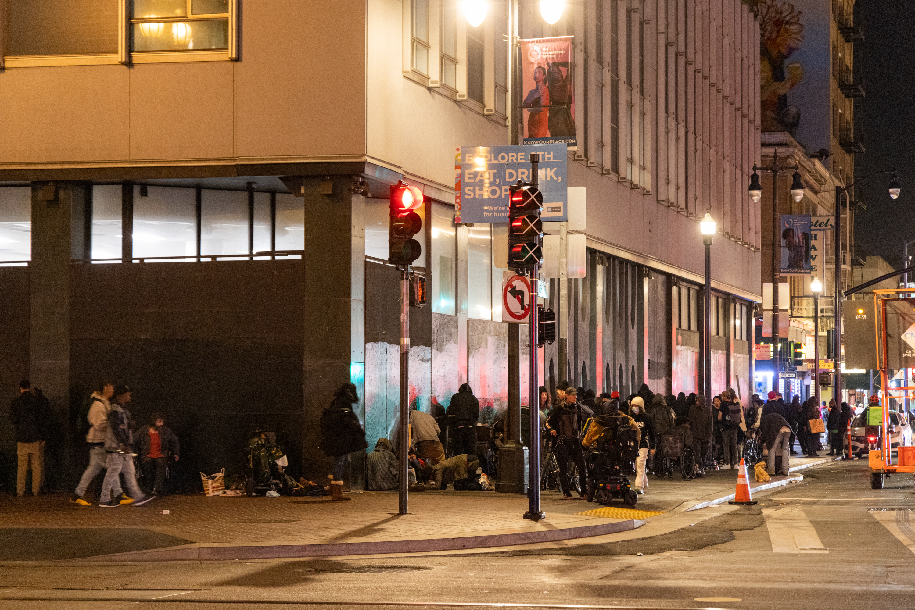 A nighttime street scene shows a large group of people gathered on the sidewalk near a busy intersection, with various signs and storefronts visible in the background.