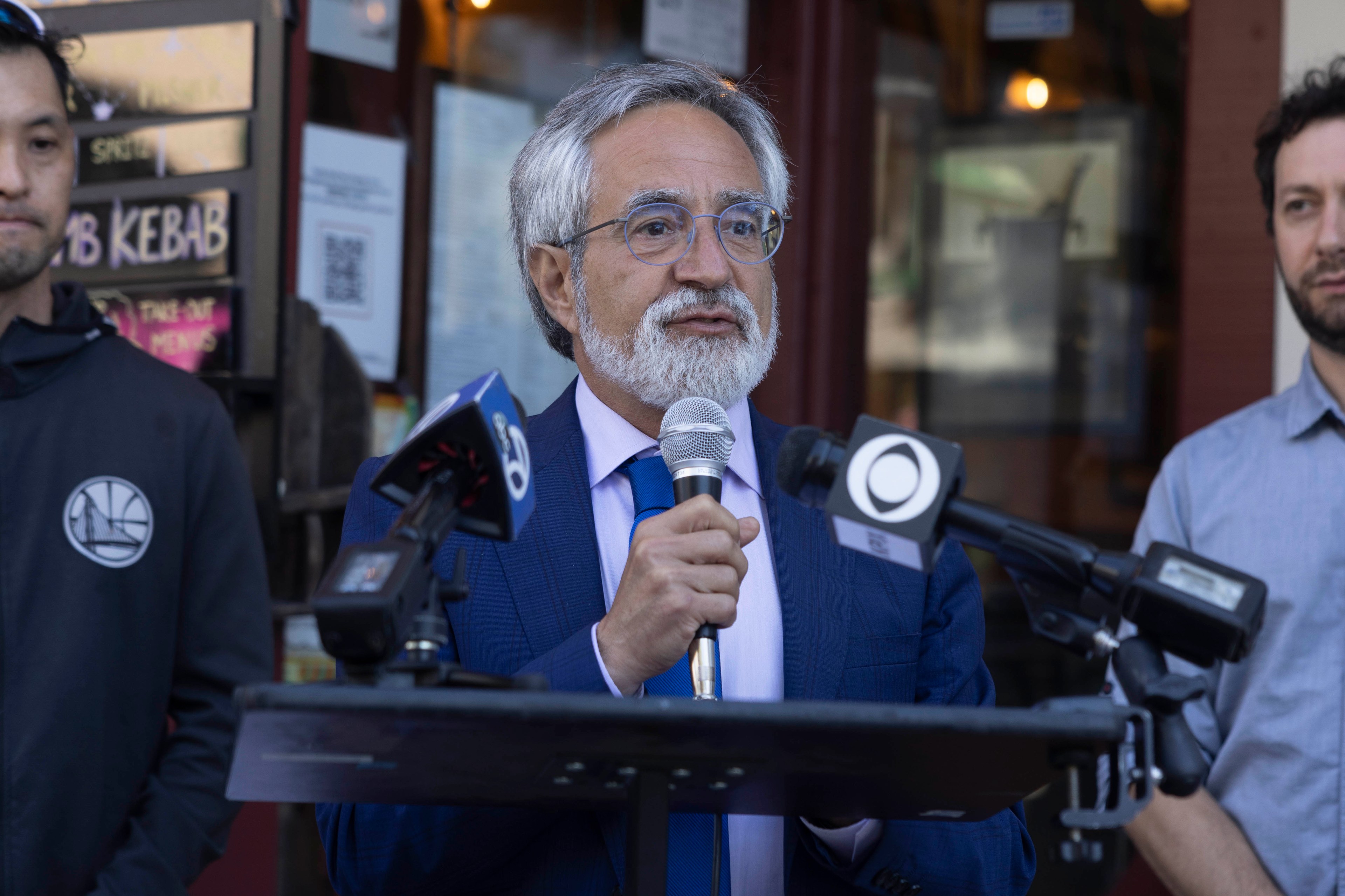 A man with gray hair and glasses, dressed in a blue suit, speaks into a microphone at a podium surrounded by microphones; two men stand nearby.