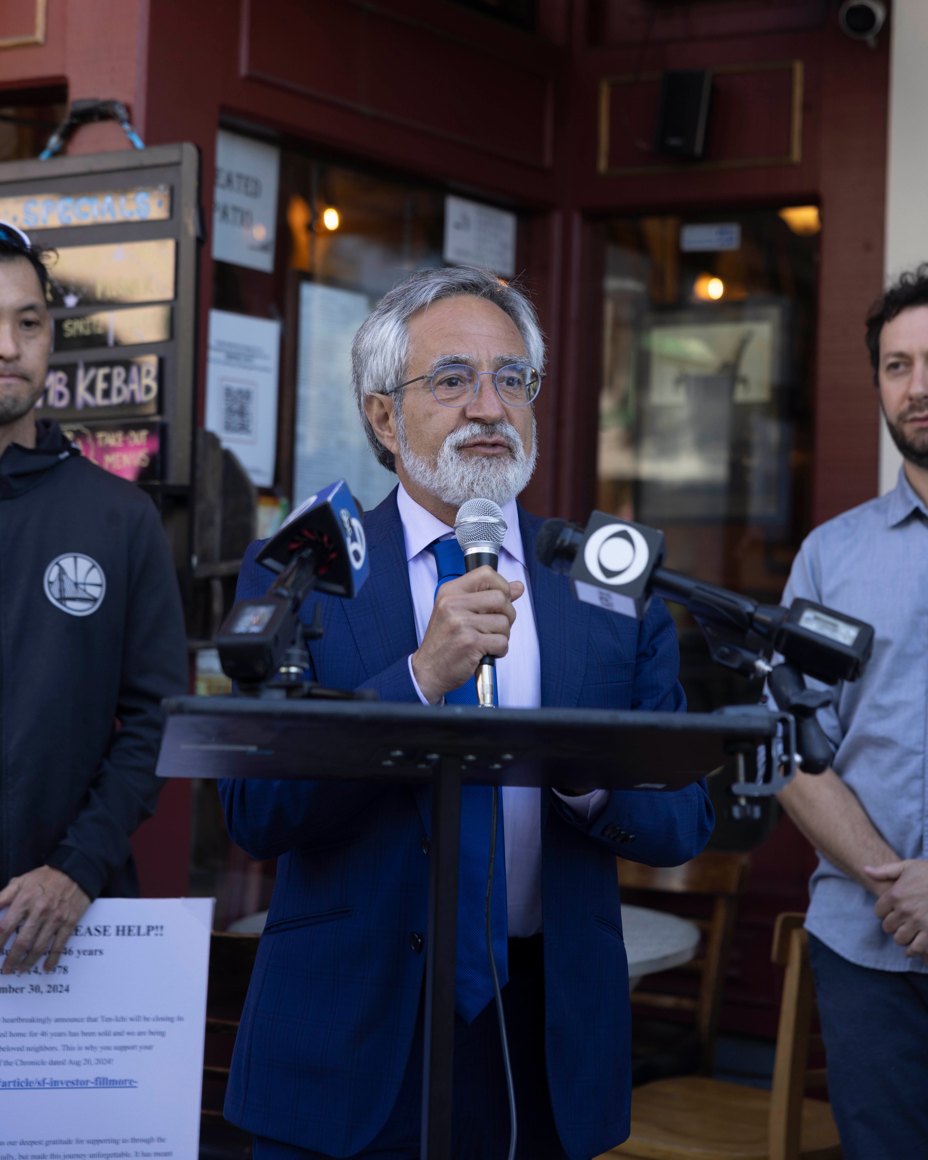 A man with gray hair and glasses, dressed in a blue suit, speaks into a microphone at a podium surrounded by microphones; two men stand nearby.