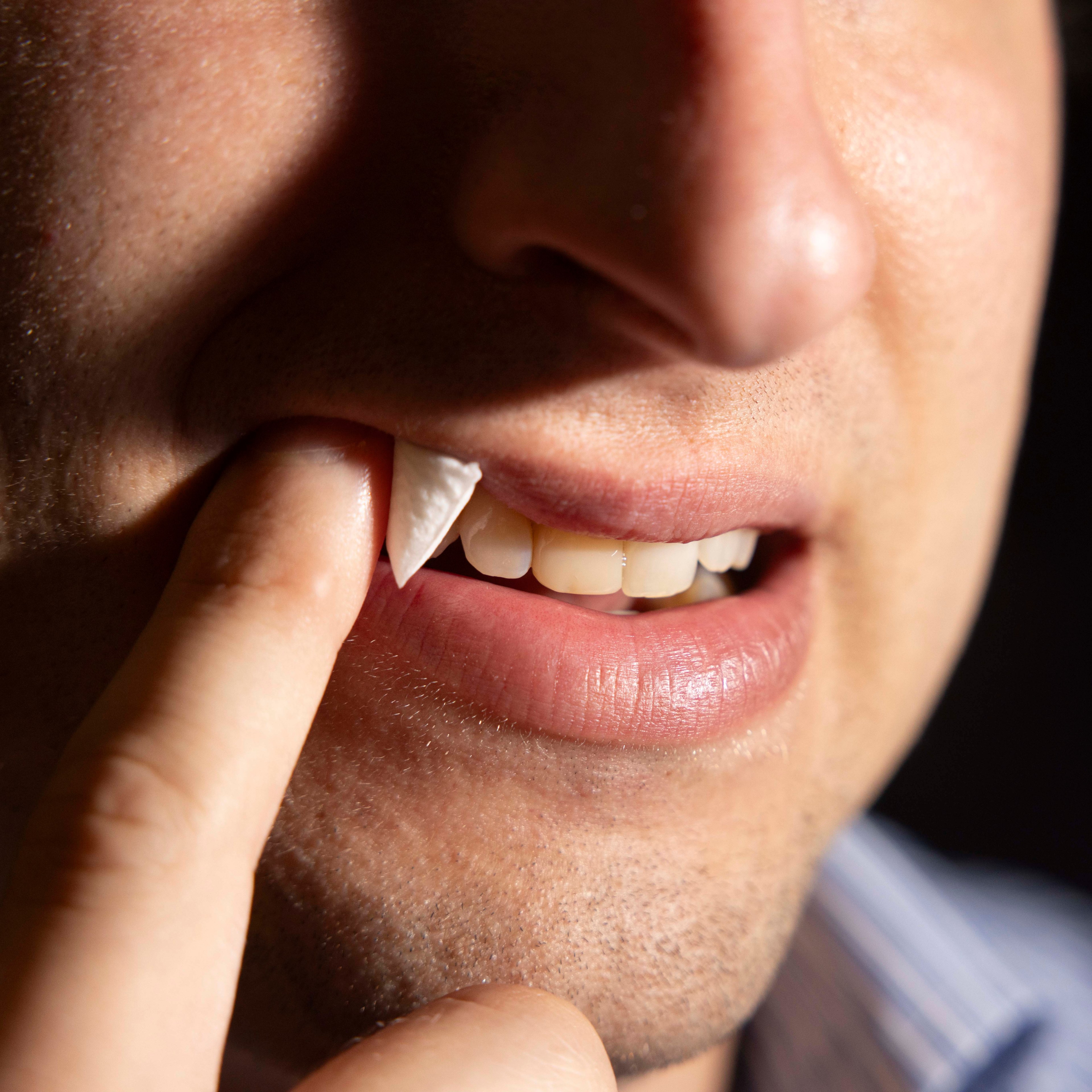 A close-up shows a person pushing a piece of white paper between their teeth and lip with their finger, focusing on their half-smiling mouth and clean teeth.