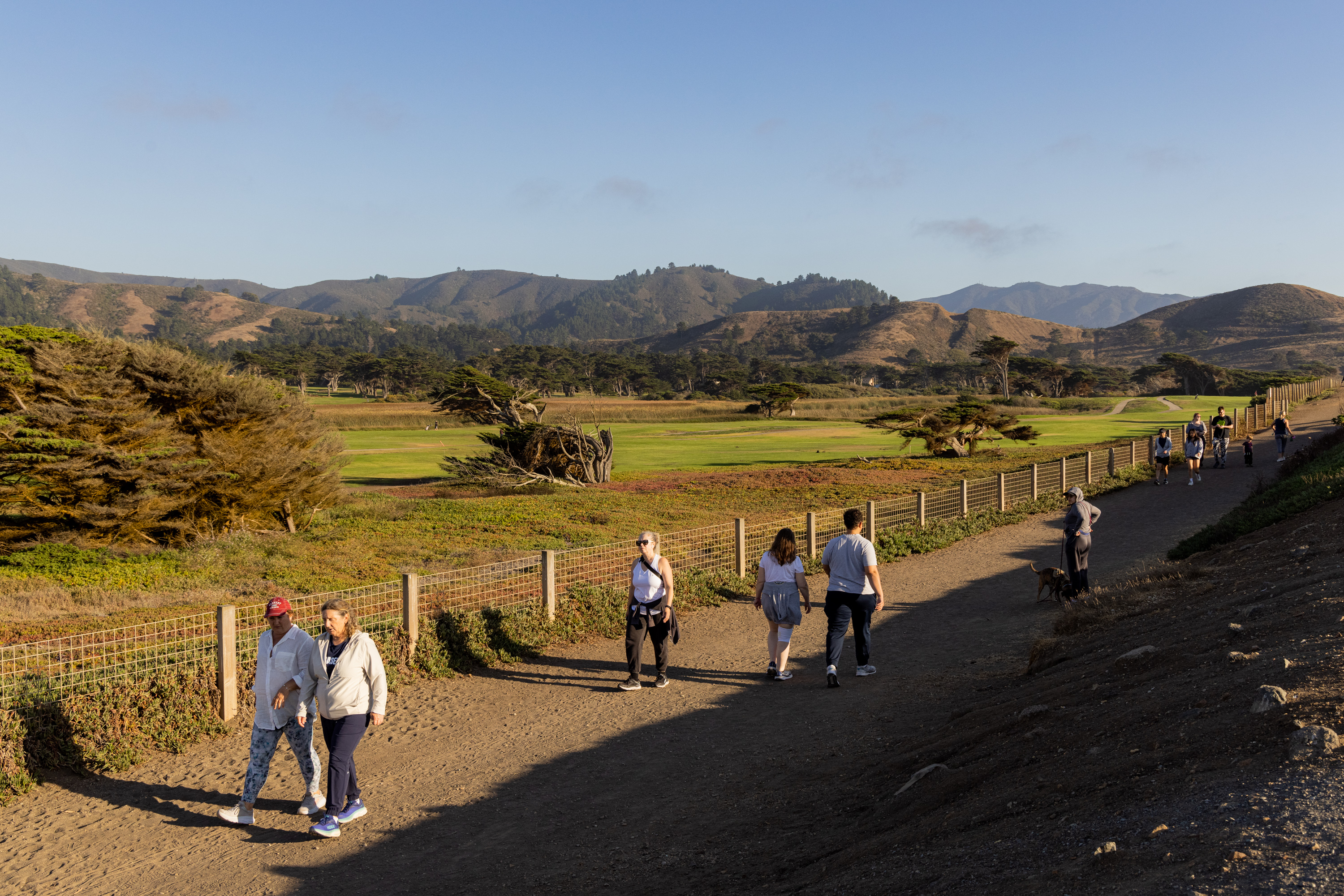 People walk along a dirt path beside a fenced grassy field, with rolling hills and trees in the background under a clear blue sky.