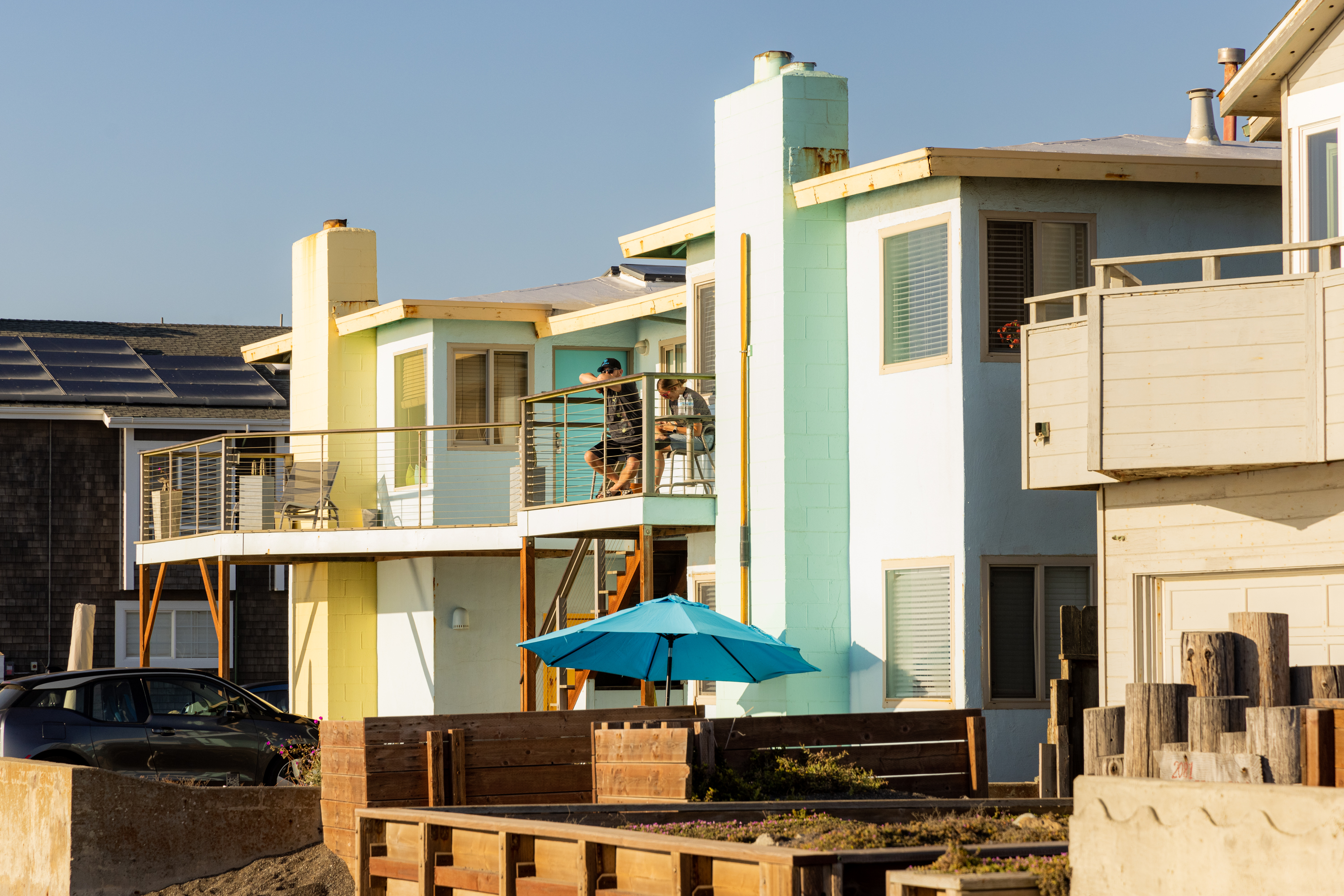 There are pastel-colored beach houses with balconies and a blue umbrella outside. People are sitting on one of the balconies, enjoying the sunny weather.