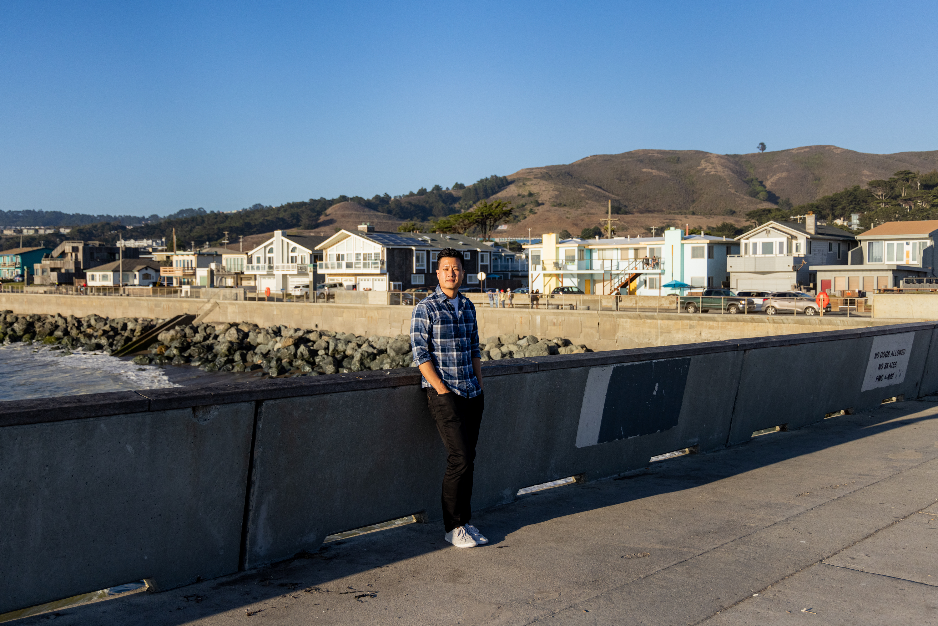 A person in a plaid shirt stands on a concrete pier beside the ocean, with houses and hilly terrain in the background under a clear blue sky.