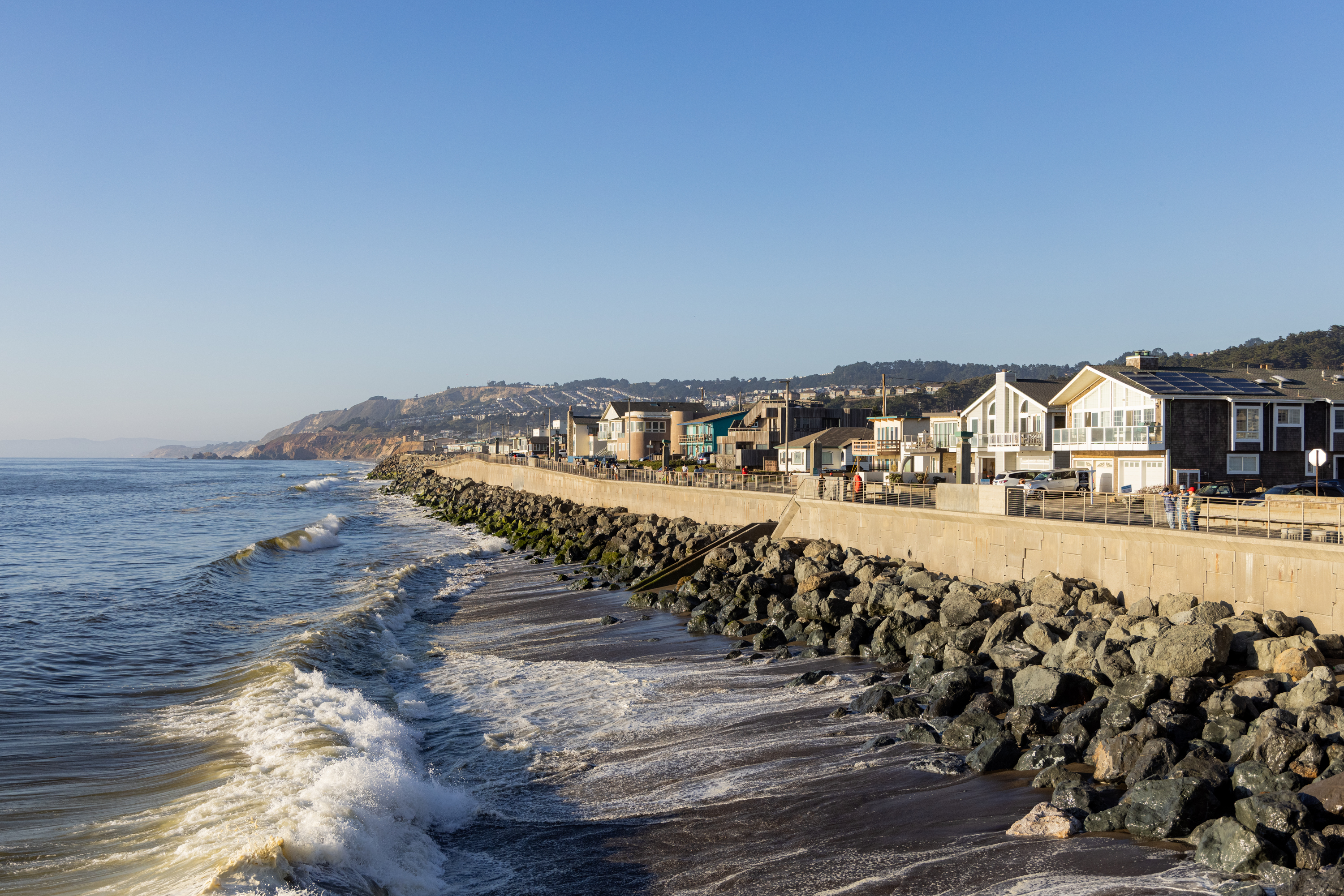 The image shows a coastal scene with waves crashing against a rocky shoreline. Above the shoreline, there is a seawall, behind which is a row of houses.