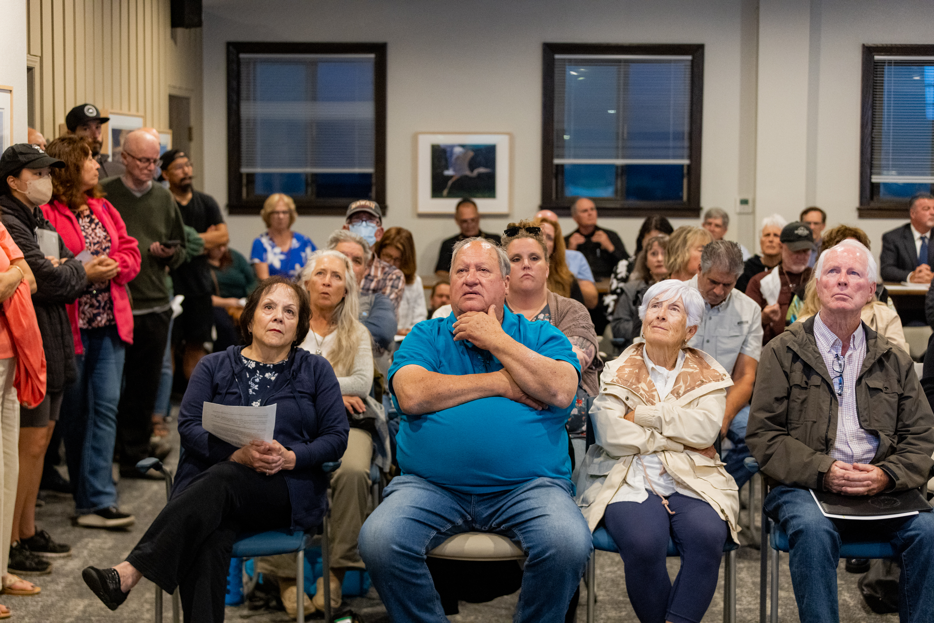 A group of people, seated and standing, gather in a room, attentively facing forward. Some hold papers, and they appear to be at a community meeting or event.