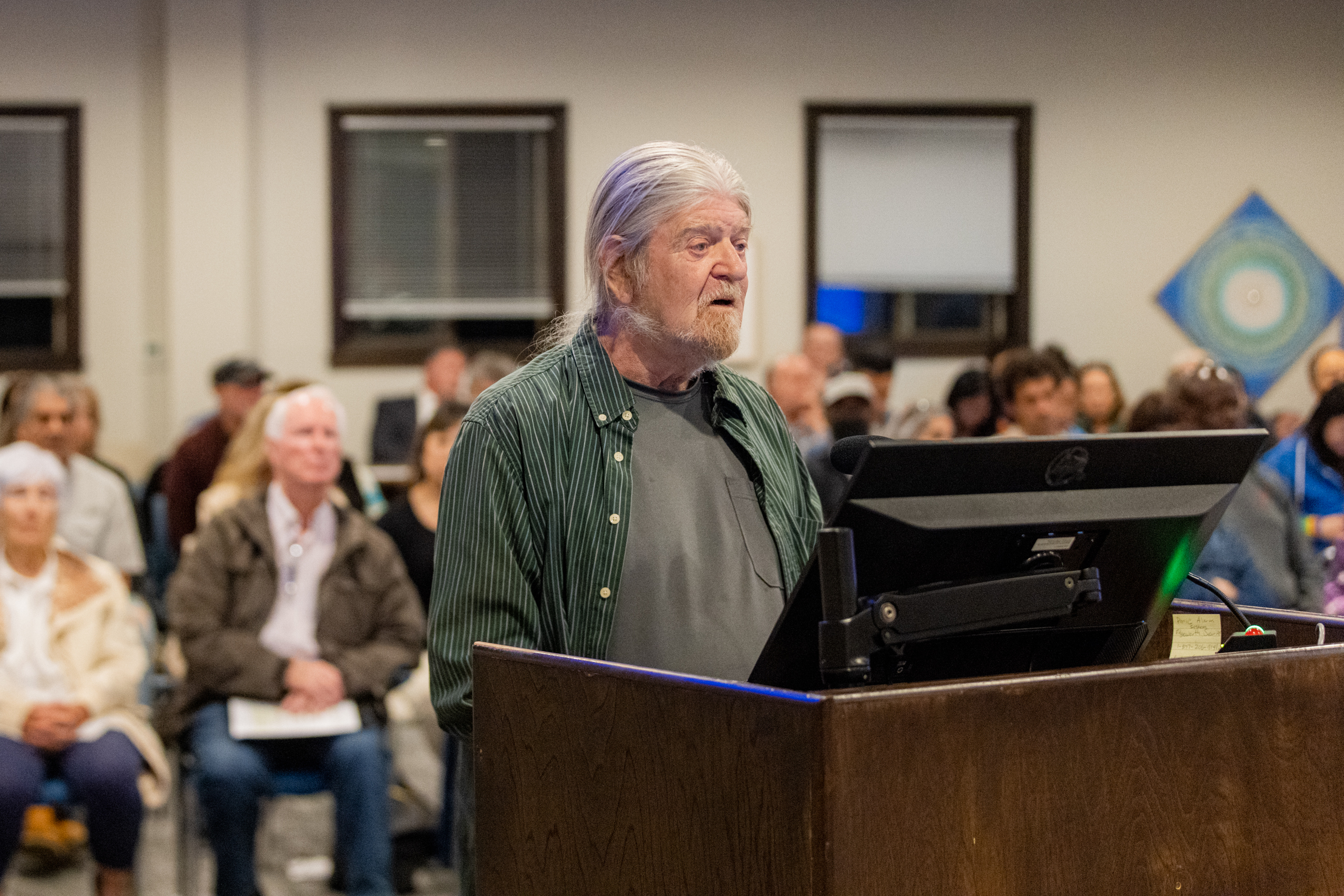 An older man with long gray hair and beard, wearing a green striped shirt, speaks at a podium in a room filled with attentive people seated in rows.