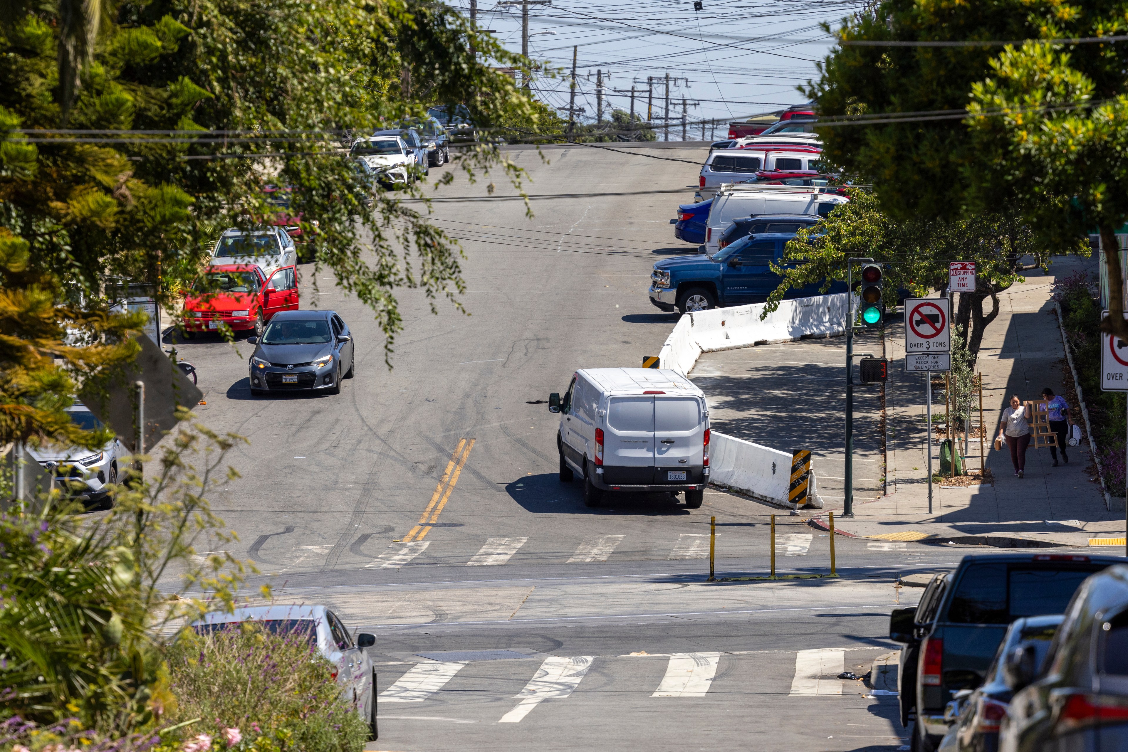 A street with cars and a white van driving uphill, surrounded by trees and parked vehicles. Two pedestrians walk on the sidewalk, and a traffic light shows green.