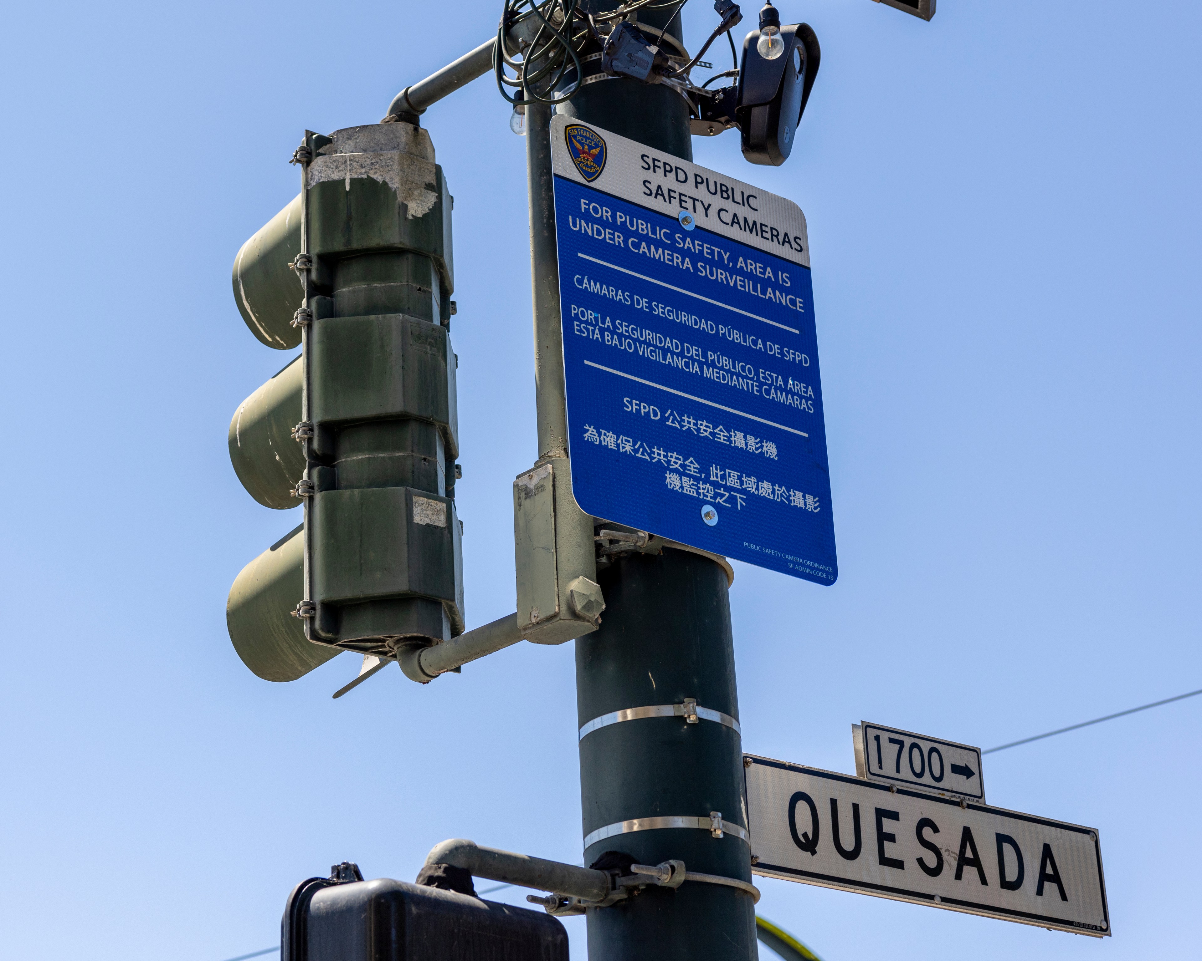 A green traffic light pole features a blue sign indicating SFPD public safety cameras. A street sign below reads &quot;1700 Quesada,&quot; against a clear blue sky.