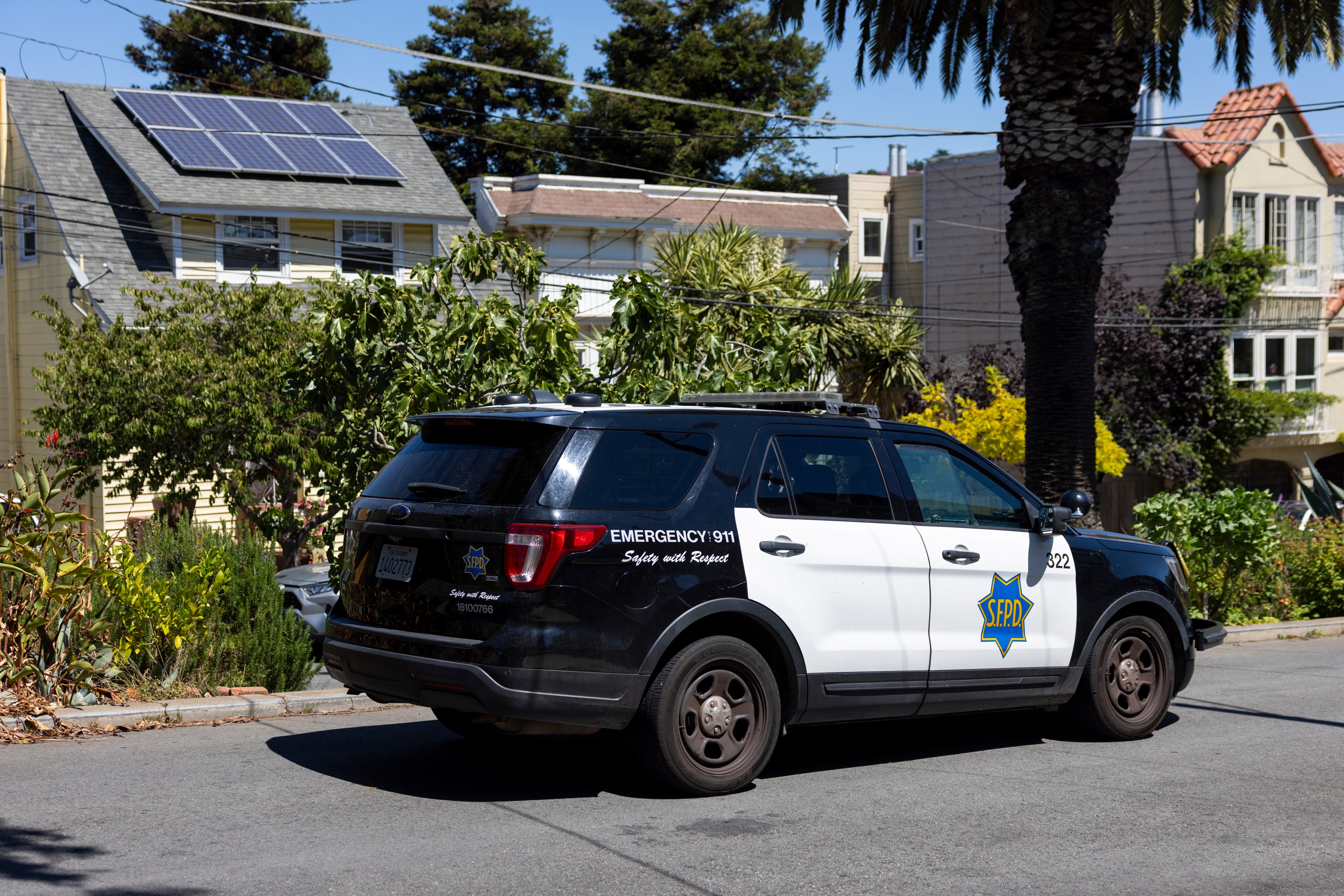 A black and white police SUV marked "SFPD" is parked on a residential street lined with trees and houses, one with solar panels on its roof.