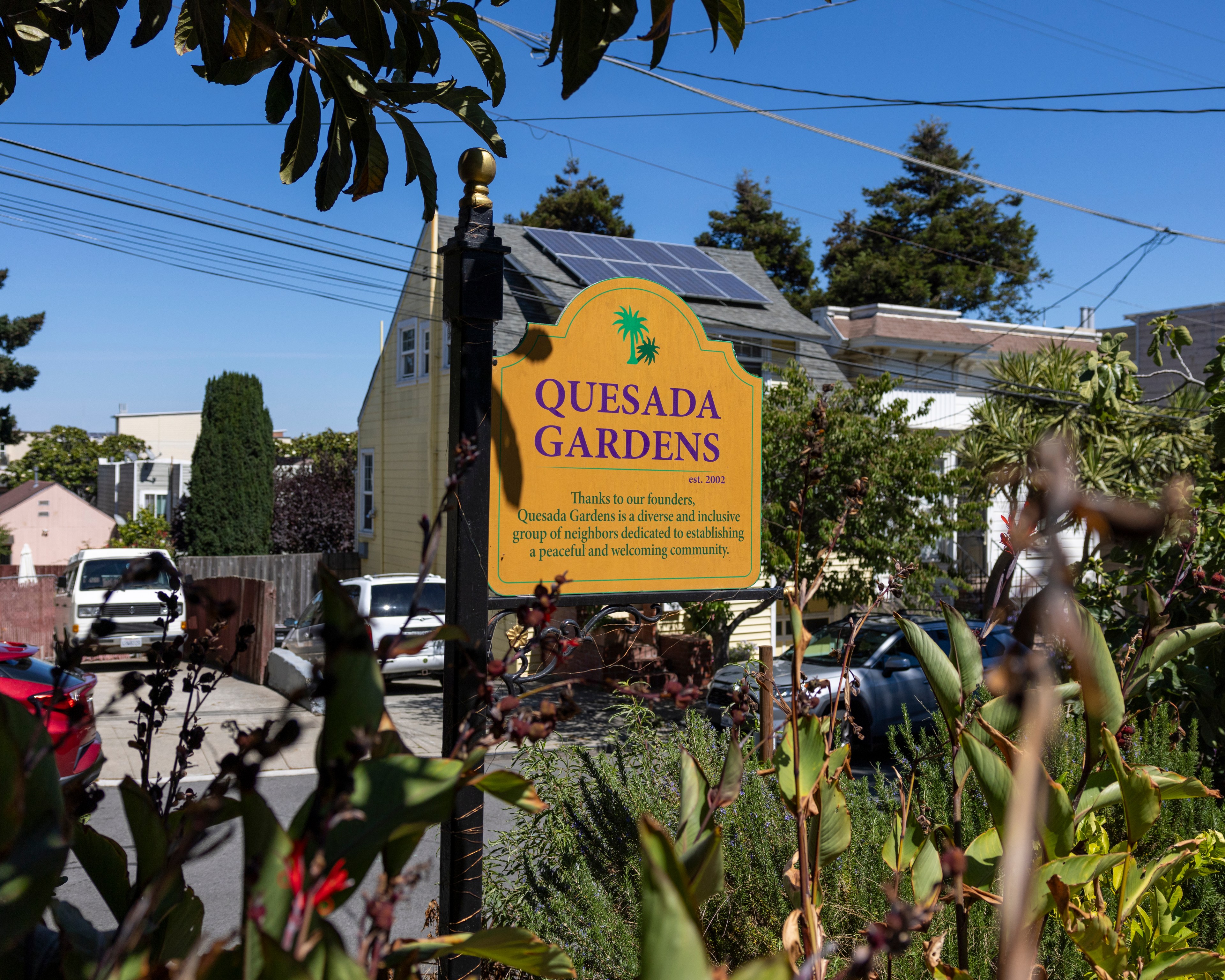 A sign in a garden reads &quot;Quesada Gardens,&quot; thanking founders and describing the community as diverse and inclusive. There are plants, houses, and cars in the background.