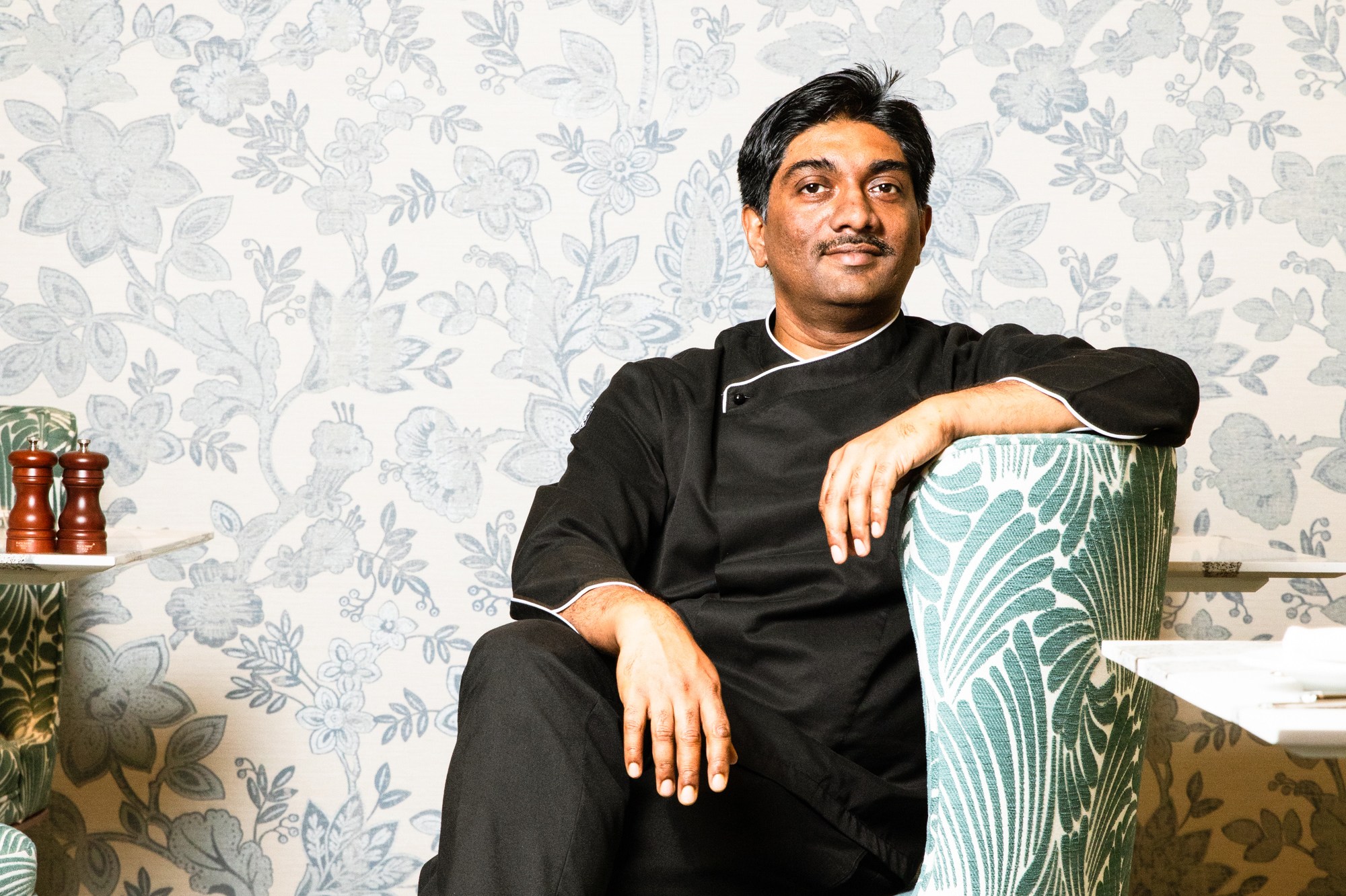 A man in black attire sits relaxed in a green-patterned armchair against a floral wallpaper backdrop, with his legs crossed and a confident expression.