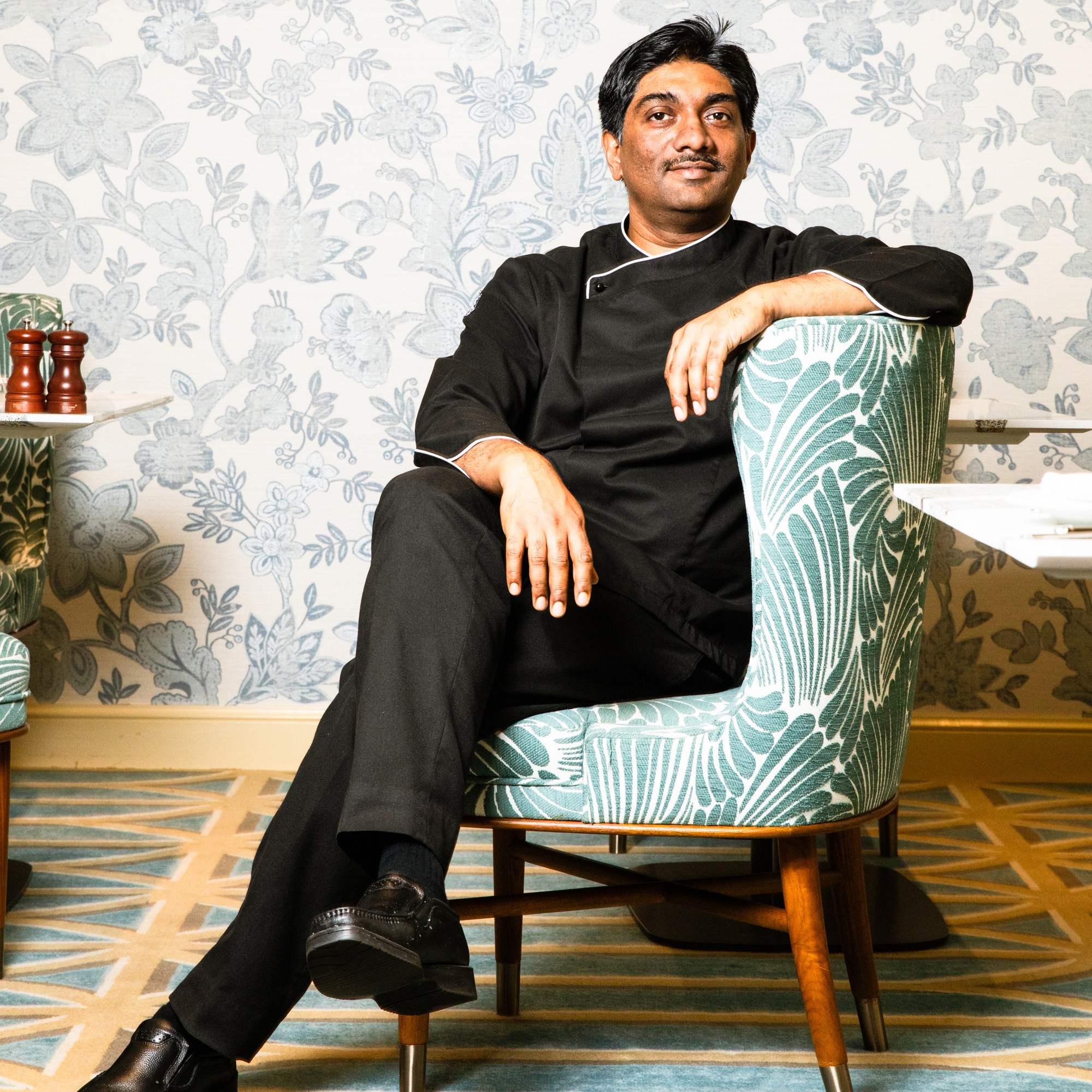 A man in black attire sits relaxed in a green-patterned armchair against a floral wallpaper backdrop, with his legs crossed and a confident expression.