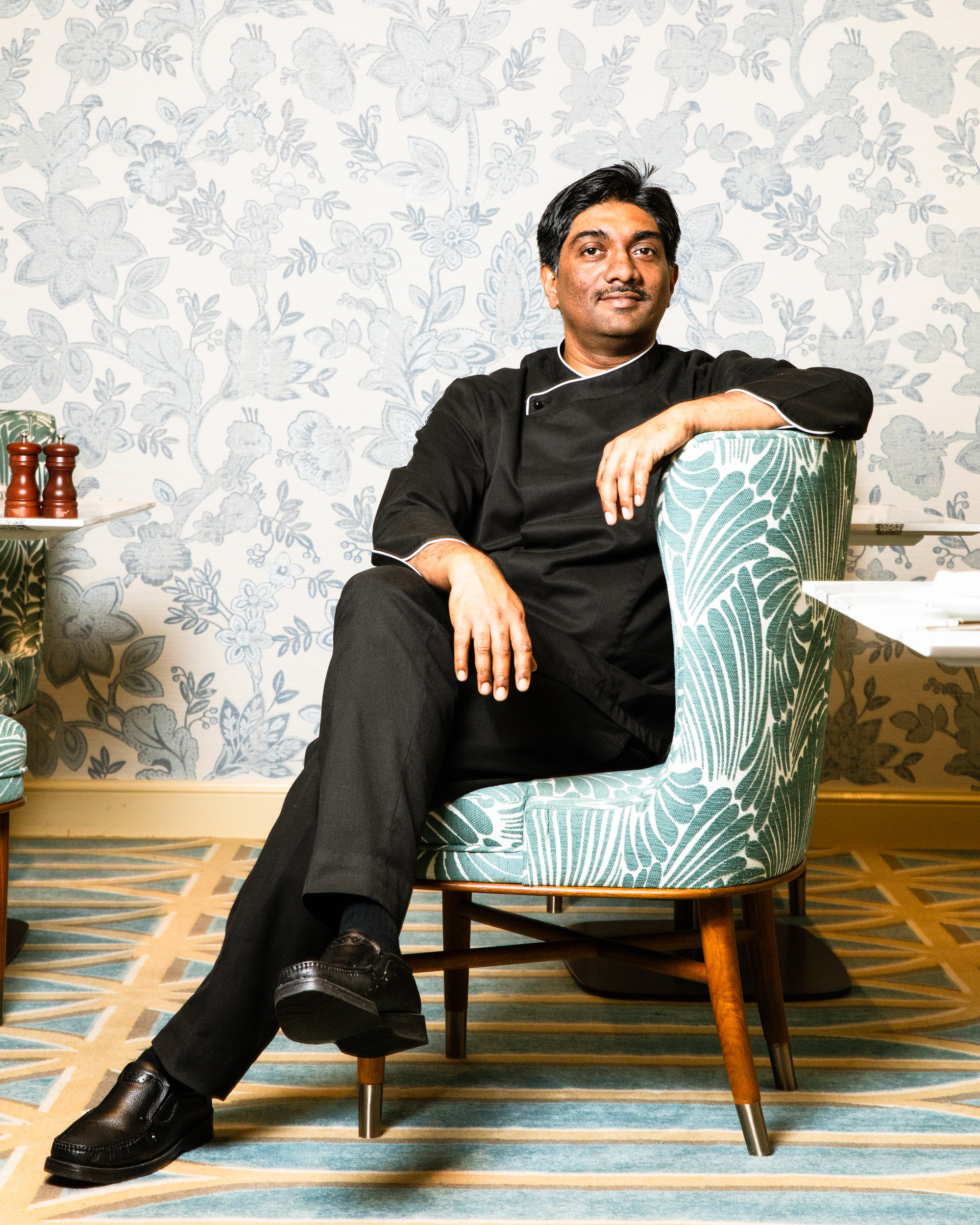 A man in black attire sits relaxed in a green-patterned armchair against a floral wallpaper backdrop, with his legs crossed and a confident expression.