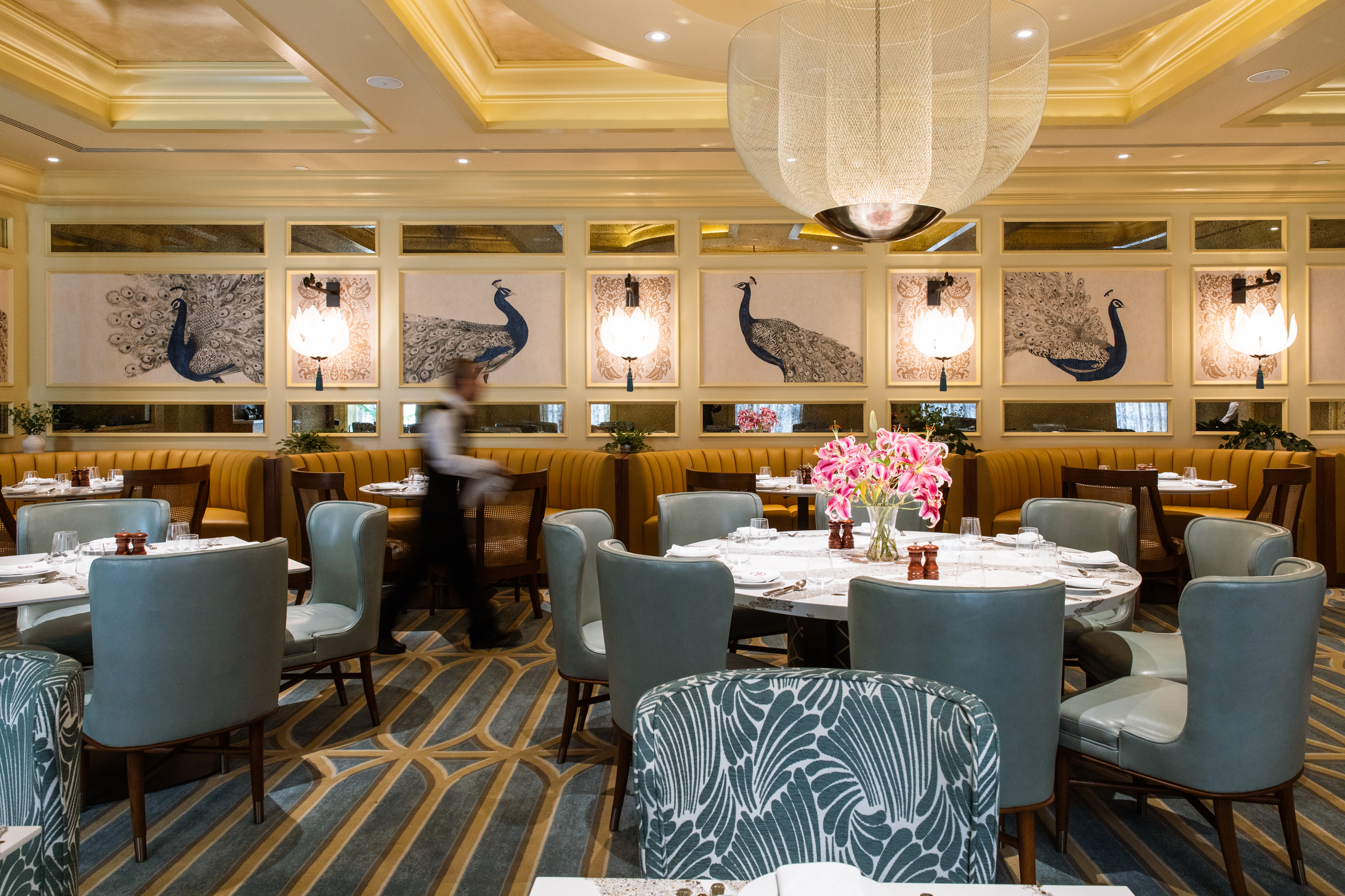 The image shows an elegant restaurant with teal chairs, yellow booths, and peacock art on the walls. A waiter walks by as pink flowers adorn the tables.