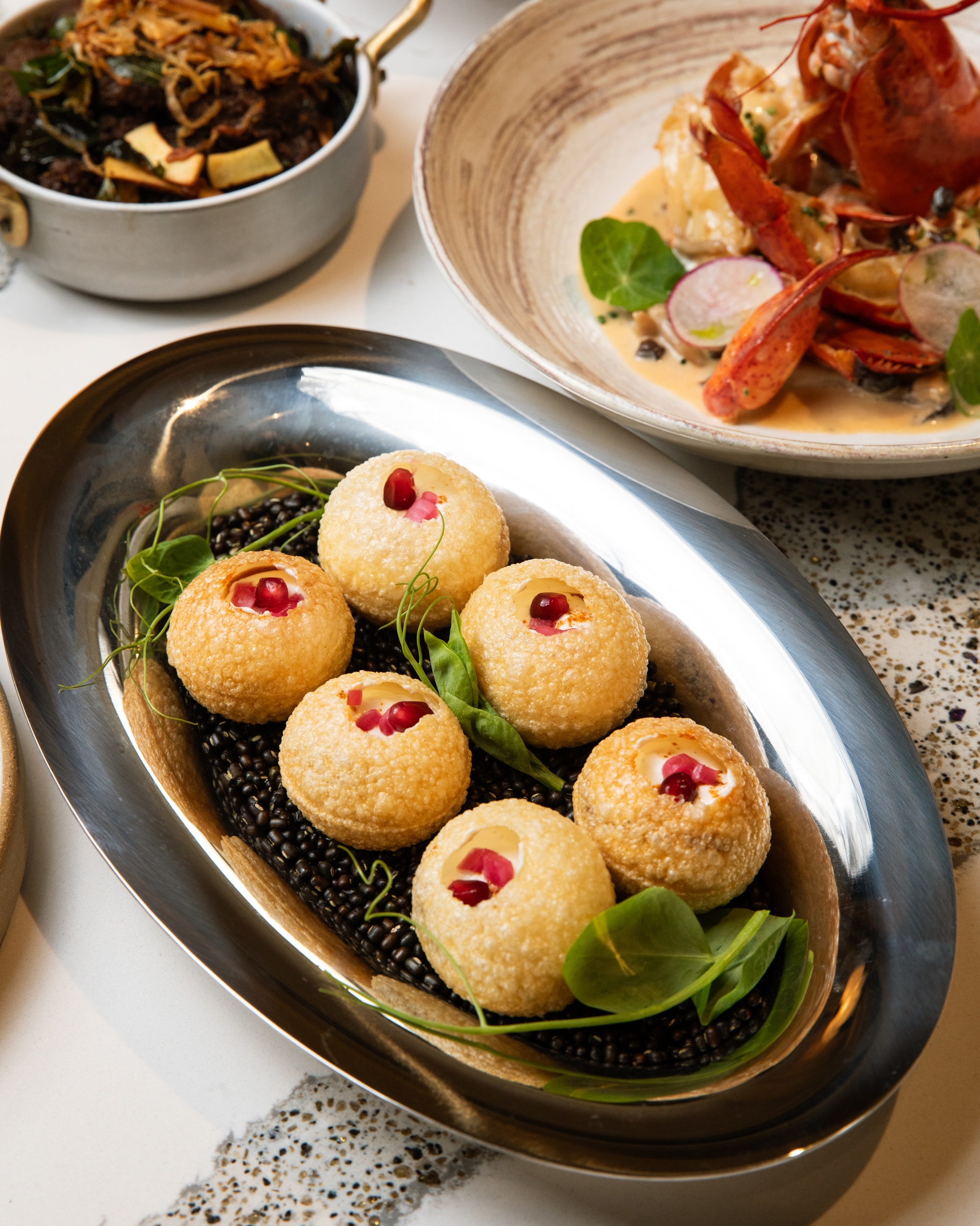 The image features a silver tray with six round, hollow, crispy puffed balls garnished with pink fillings and greenery, along with two other dishes in the background.