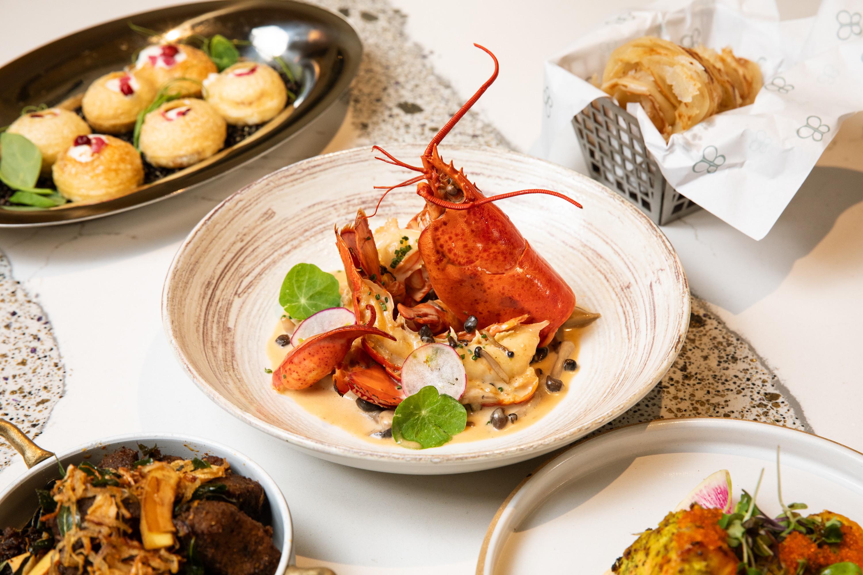 A table set with various gourmet dishes including a lobster main course with garnish, a basket of bread, a plate of puff pastries, and another dish with meat and greens.