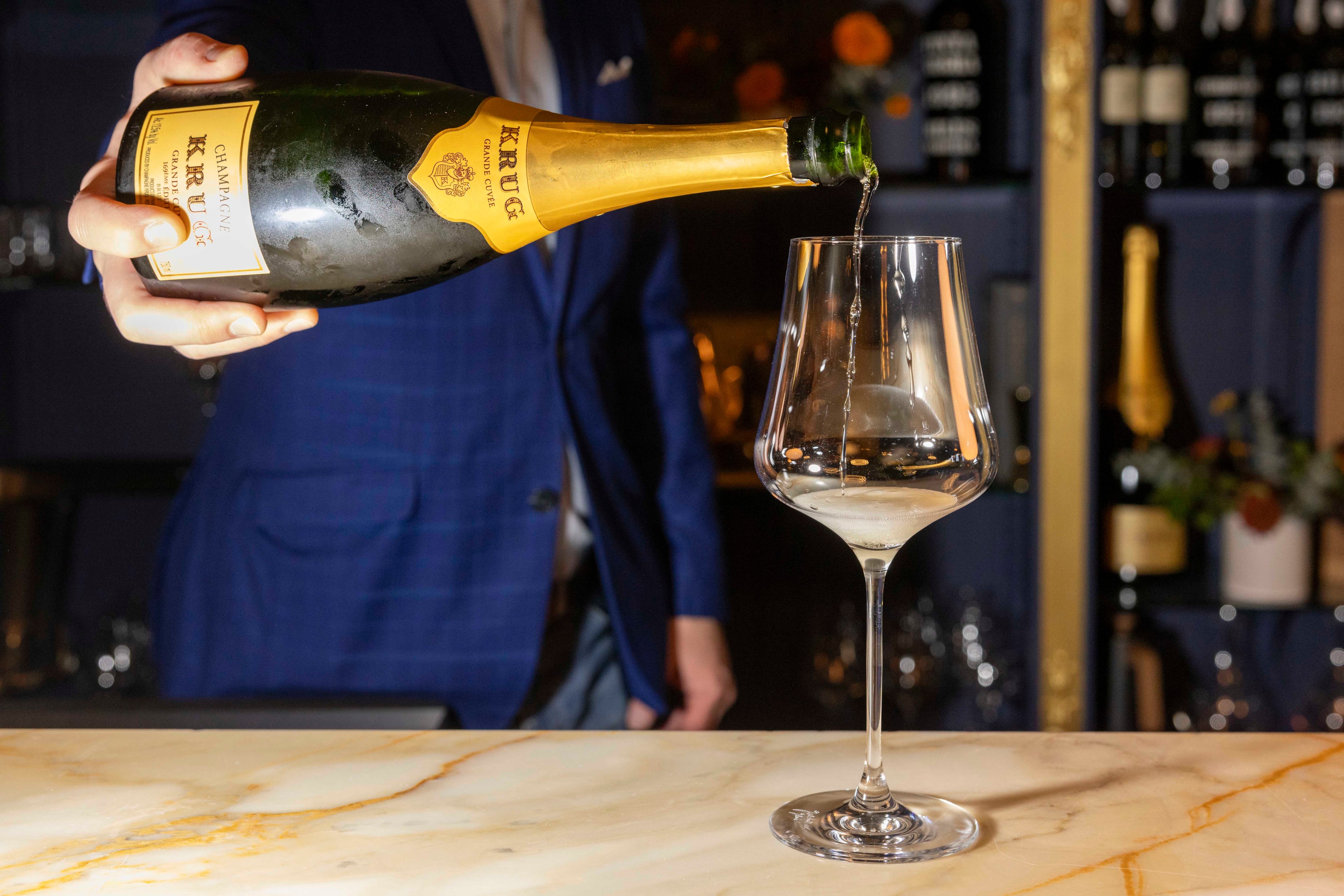 A person in a blue jacket pours champagne from a bottle labeled "Krug" into a wine glass on a marble surface, with a bar shelf in the background.